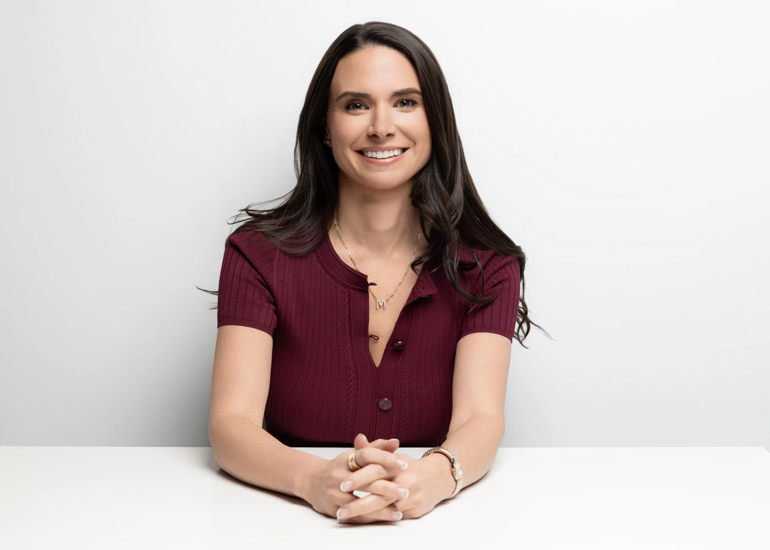 A woman with long dark hair smiling, wearing a maroon short-sleeve button-up top, sitting at a white table against a plain white background.