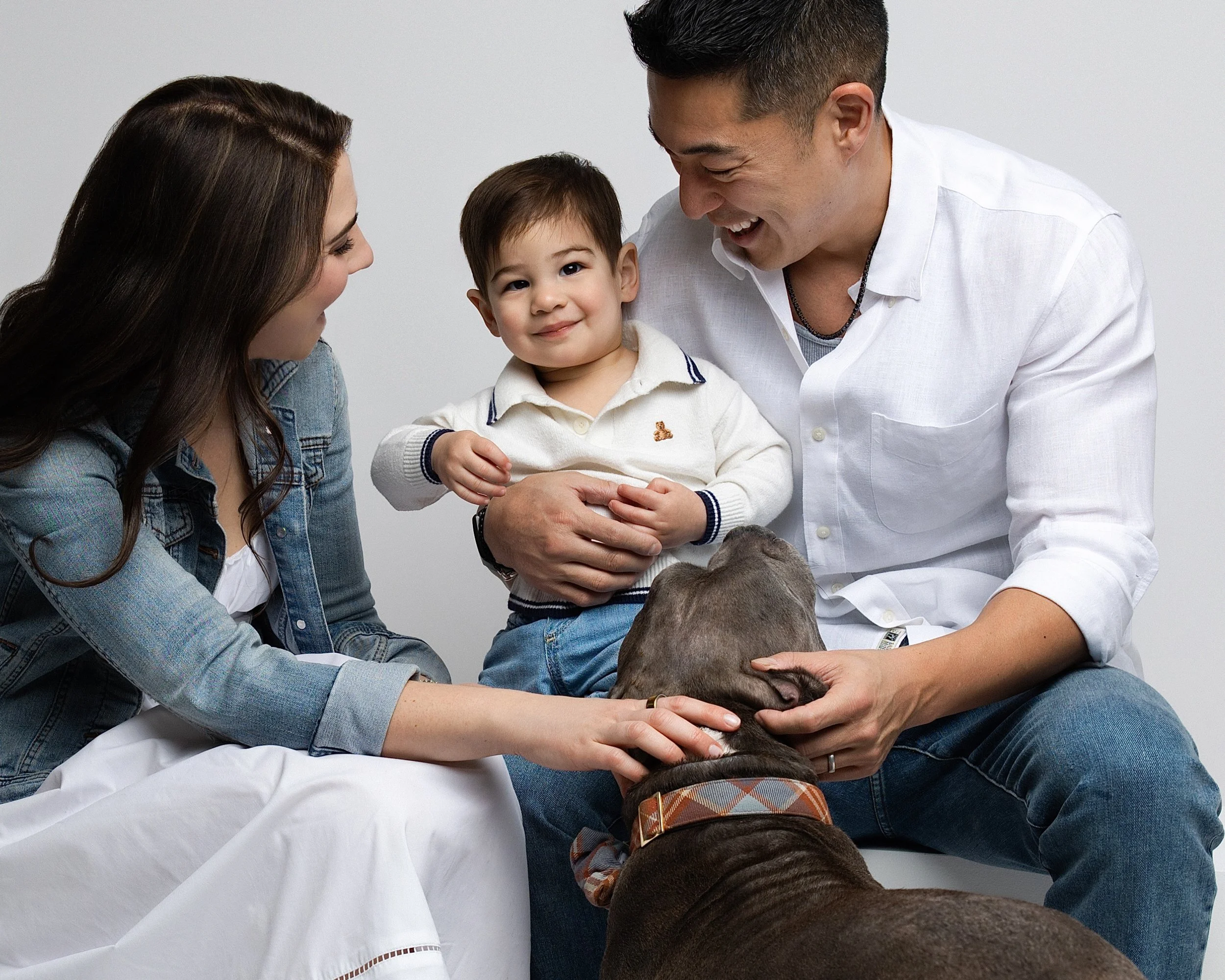 A family of three, including a woman with long brown hair, a man with short dark hair, and a young boy, is sitting together interacting with a large brown dog. The woman and man are smiling and petting the dog, while the boy, sitting between them on 