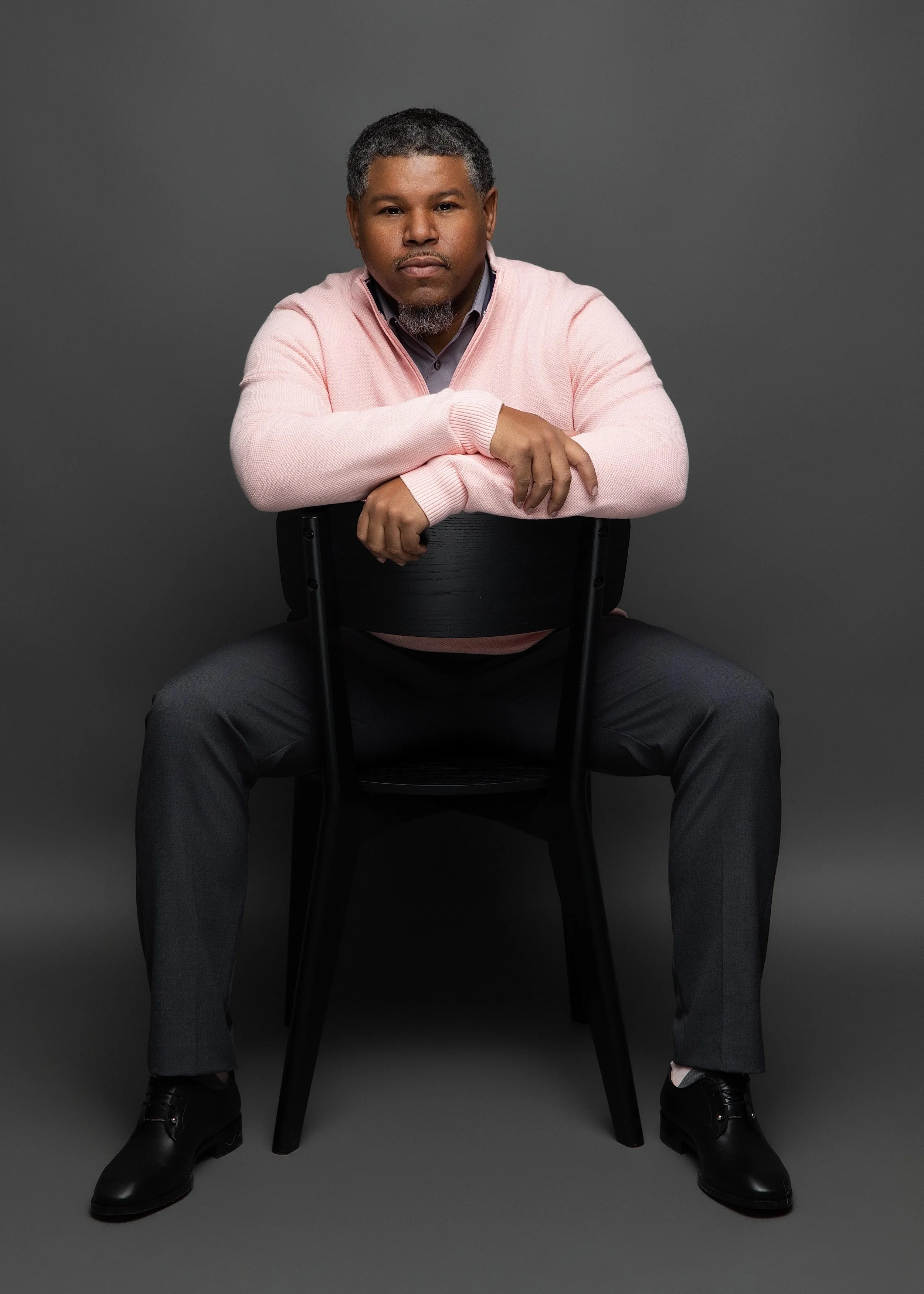 Portrait of a man sitting on a black chair against a gray background, wearing a pink sweater, gray pants, and black shoes, with short curly hair and a beard.