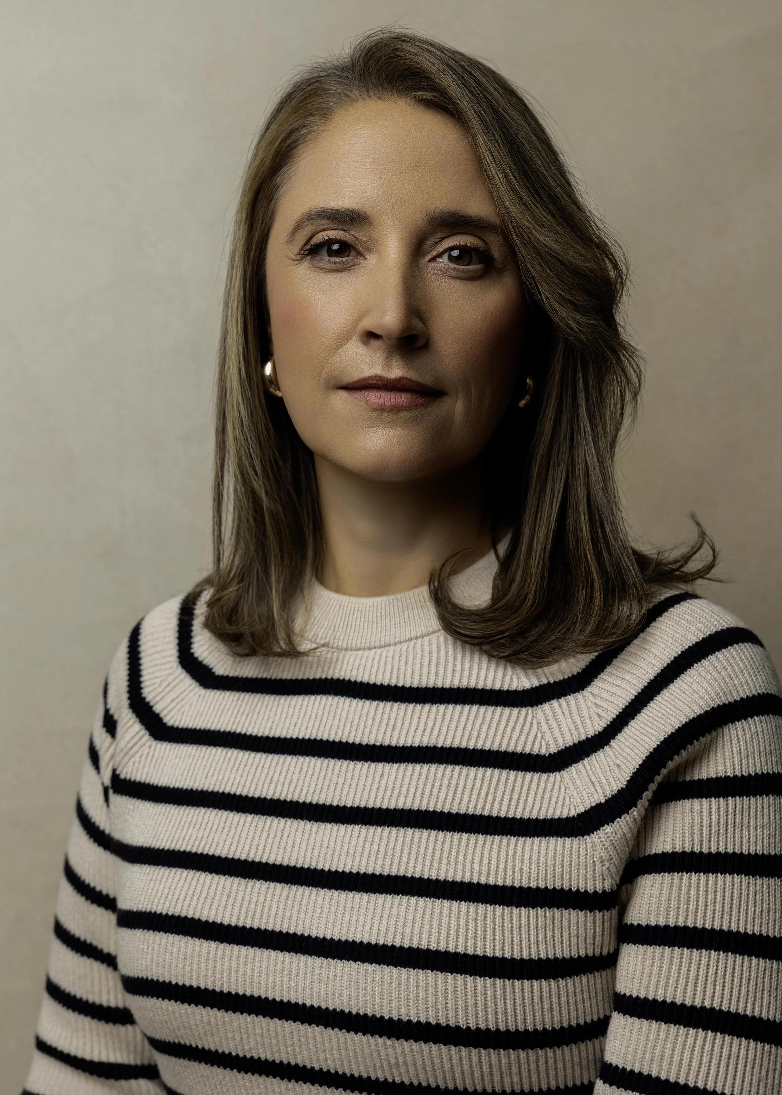 A woman with shoulder-length brown hair, wearing a cream and black striped sweater, gold hoop earrings, and minimal makeup, standing against a beige background.