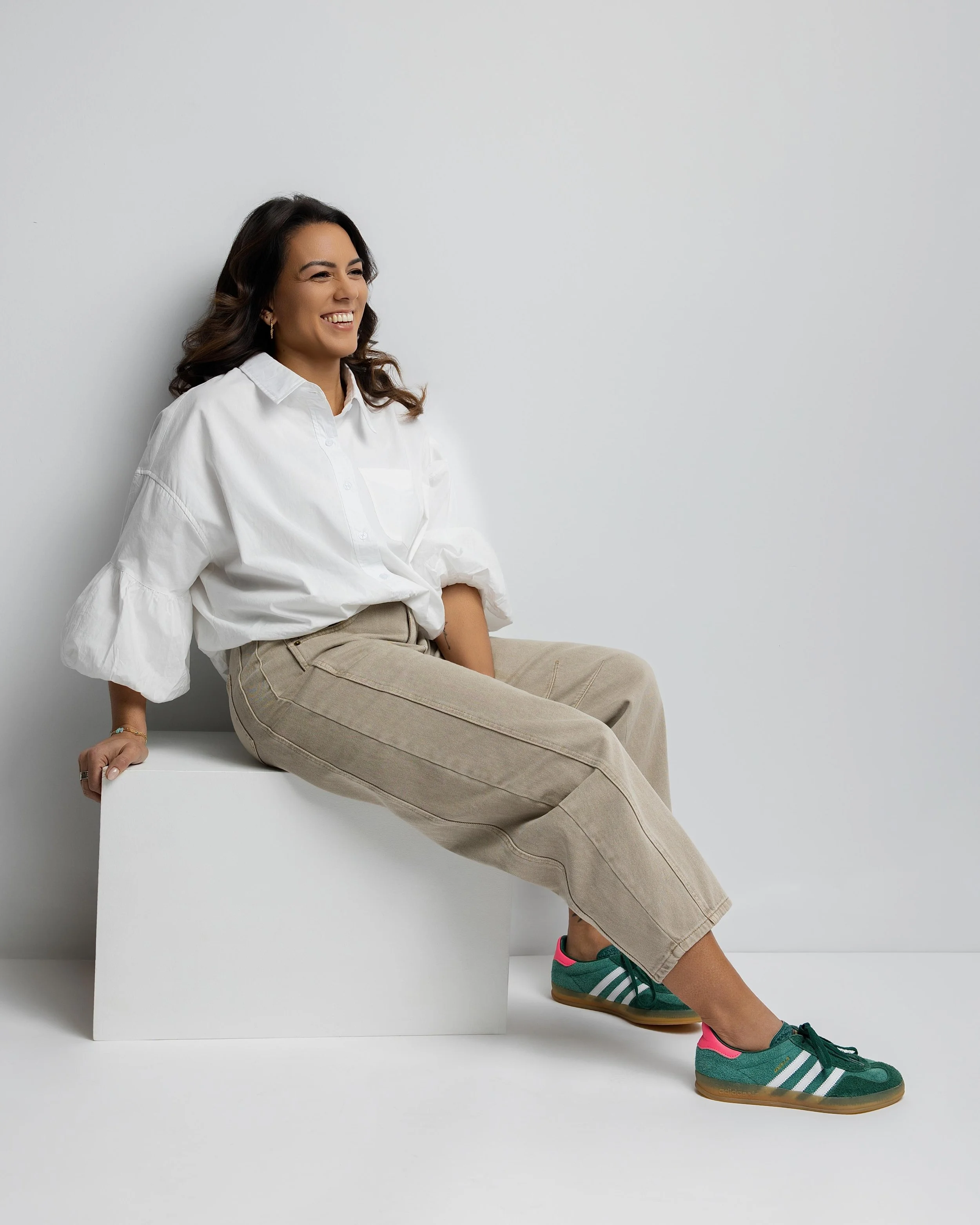 Woman sitting on a white block against a plain background, smiling and looking to her left, wearing a white button-up shirt, beige pants, and teal and pink sneakers.
