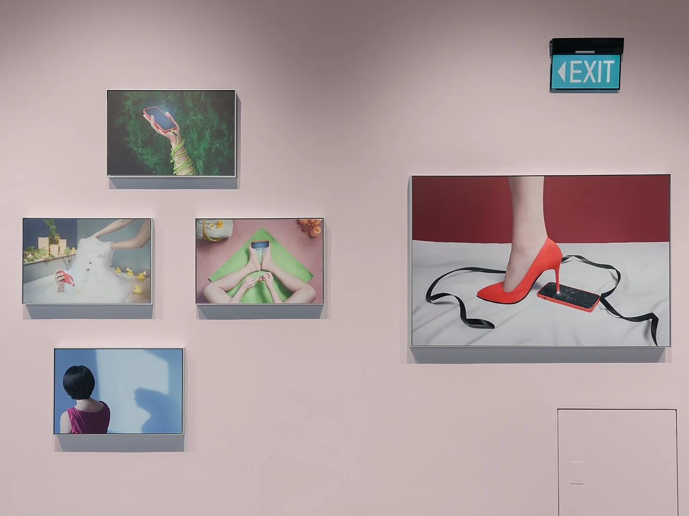 Installation of A Modern Day Affair exhibition under Our Own Frame, curated by DECK Photography Art Centre. Pale pink gallery wall with five framed photographs; the largest frame shows a red high heel pressing into a cracked smartphone.