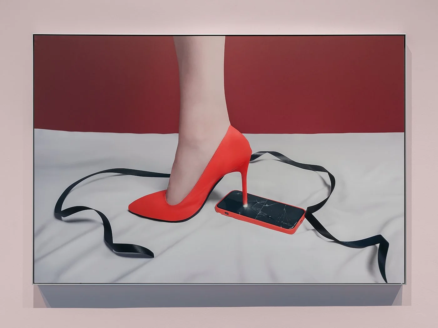 Framed photograph on a pale pink wall showing a red stiletto heel pressing into a cracked-screen smartphone with a red case; a black ribbon lies in loose loops on the surface.