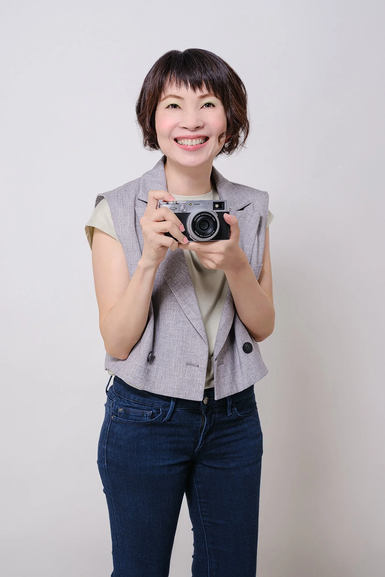 A woman smiling and holding a camera in front of a plain light gray background.