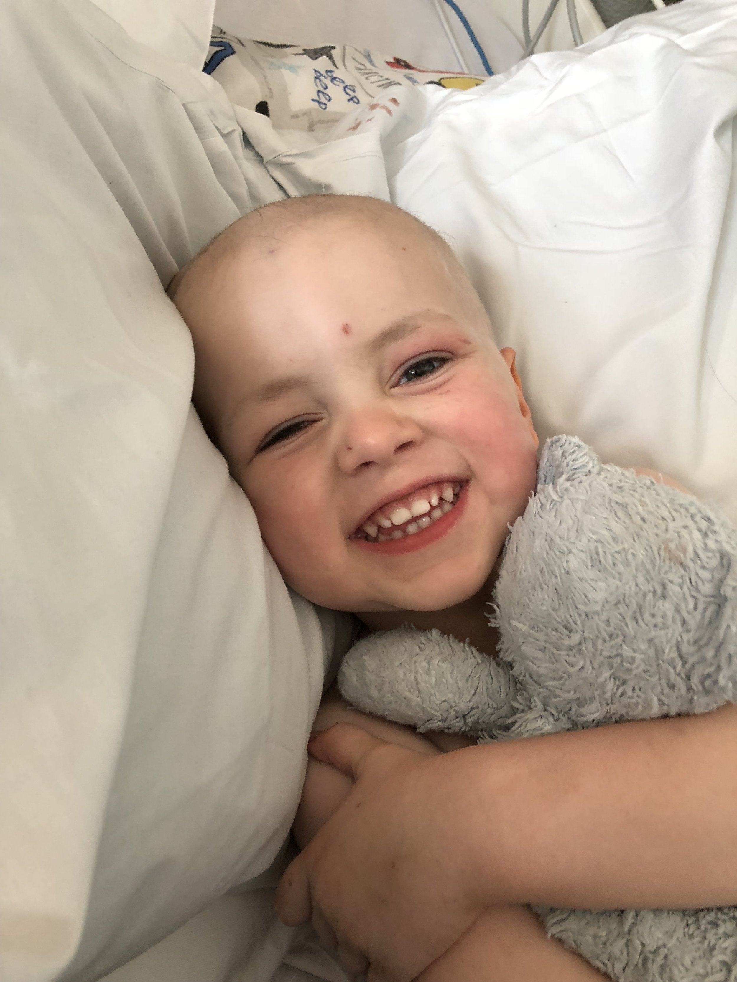 Happy young boy with no hair, smiling and lying in a hospital bed holding a gray teddy bear.