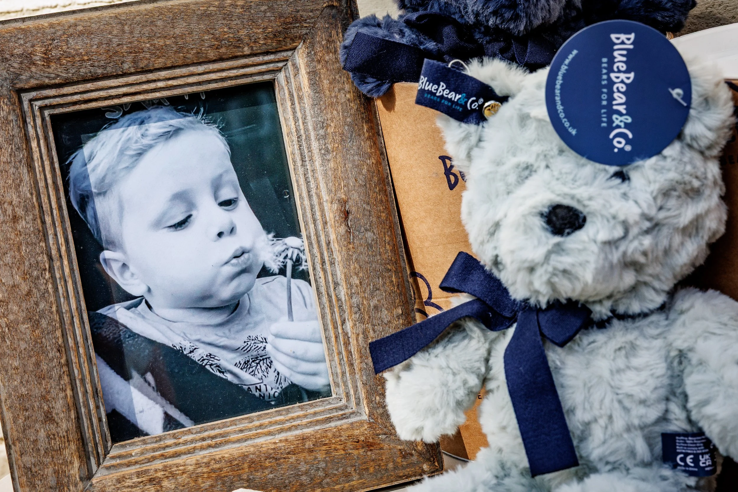 Black-and-white photo of a young boy with light hair holding a small object close to his mouth, placed in a rustic wooden frame. Next to the photo, there is a white teddy bear with a navy blue ribbon around its neck and a blue tag that reads "Blue Bear & Co."