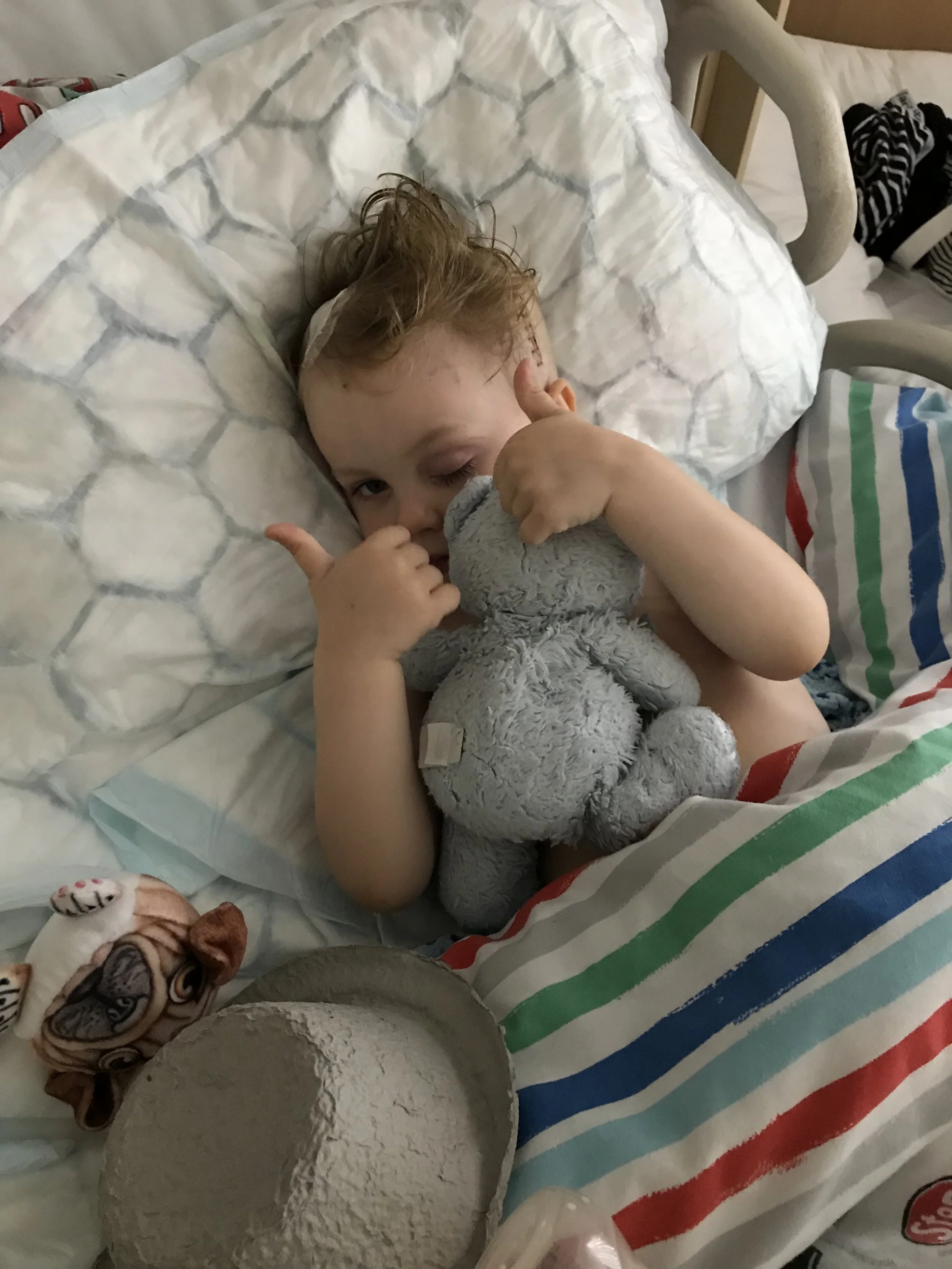 A young child with curly hair lying in a hospital bed, holding a gray stuffed animal, giving a thumbs-up, with a blanket featuring red, blue, green, and white stripes partially covering them.