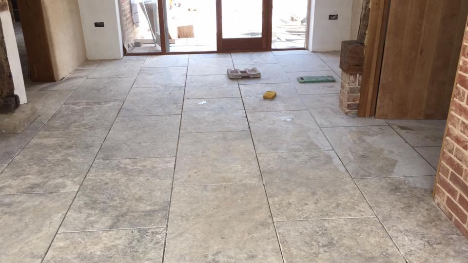 Room with gray stone tile flooring, open wooden double doors, sandpaper, sponge, and handle tools on floor, exposed brick walls.