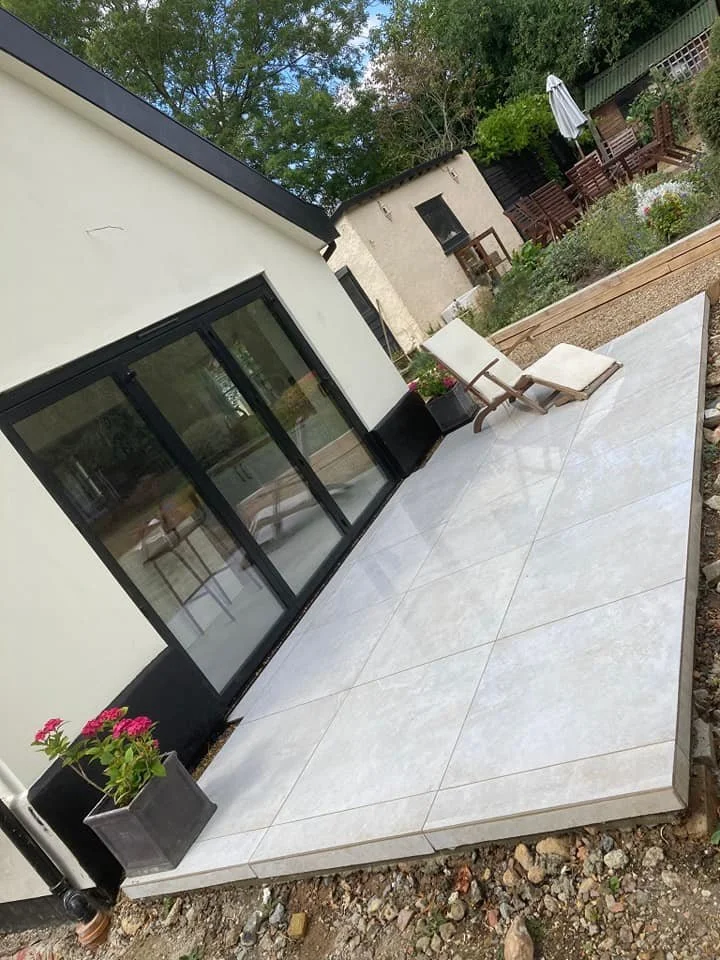Modern patio with tile flooring, lounge chairs, sliding glass doors, and a garden with outdoor furniture.