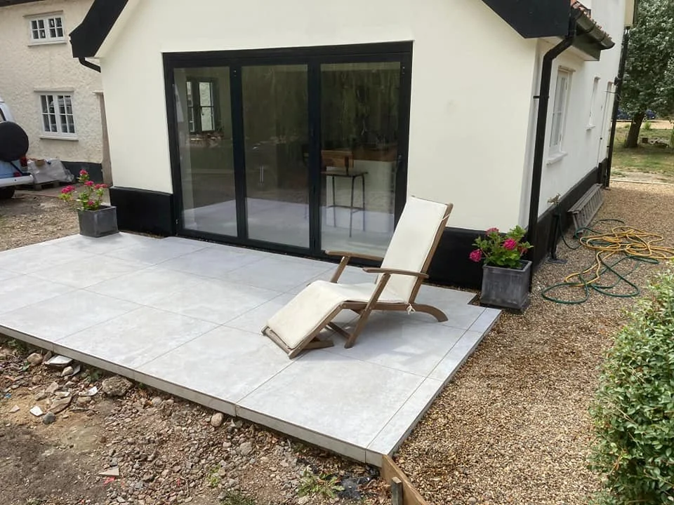 Tiled outdoor patio with a wooden deck chair, potted pink flowers, black-framed sliding glass door, and garden hose on gravel area.