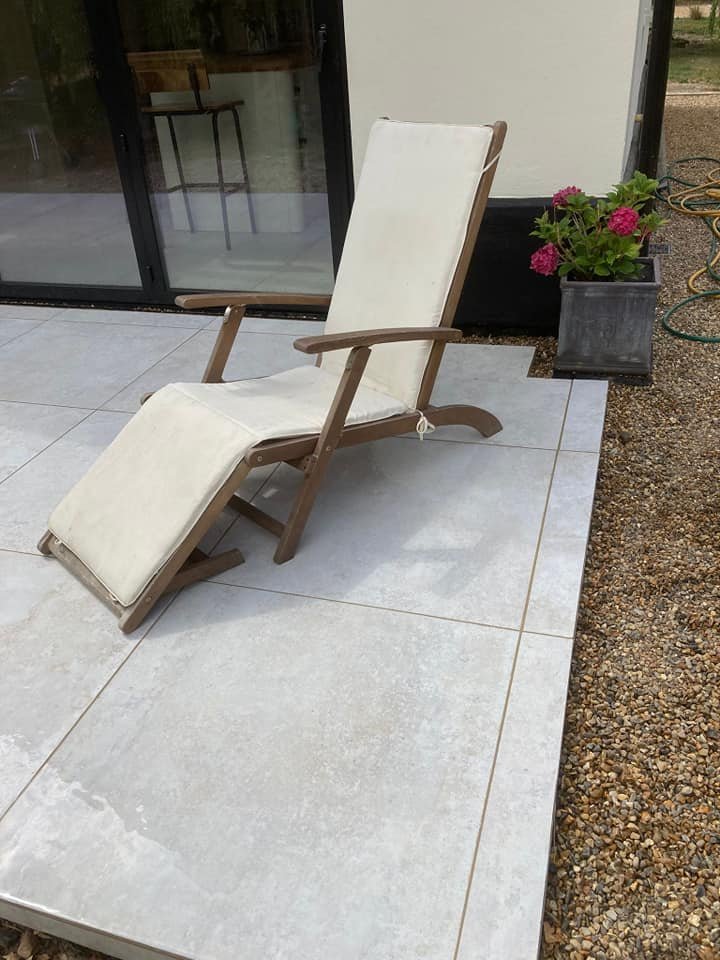 Outdoor patio scene with a wooden reclining chair and beige cushion set on light stone tiles, a potted plant with pink flowers, and a coiled garden hose.