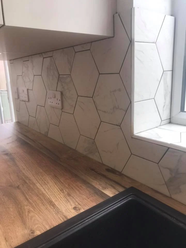 Kitchen countertop with wood finish, hexagonal marble tile backsplash, light gray hex tiles, two electrical outlets, and window sill with tiles.