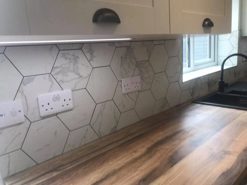 Modern kitchen countertop with wooden surface and hexagonal marble tile backsplash, featuring wall-mounted electrical outlets and under-cabinet lighting.