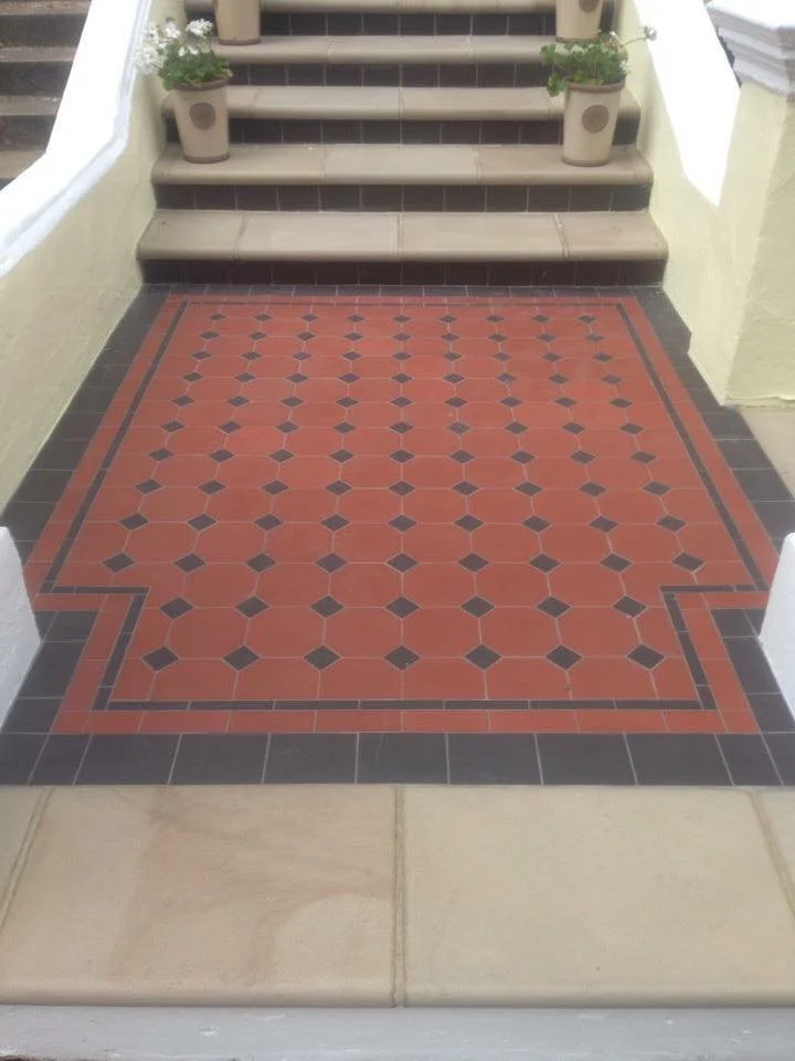 Decorative tiled entrance with red, black, and beige geometric pattern, steps, and potted plants on the sides.