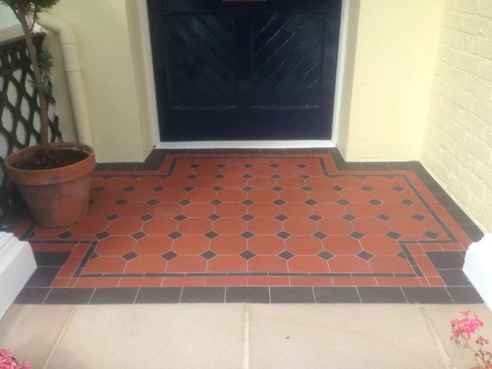 Victorian-style tiled porch with decorative pattern, potted plant, and dark door.