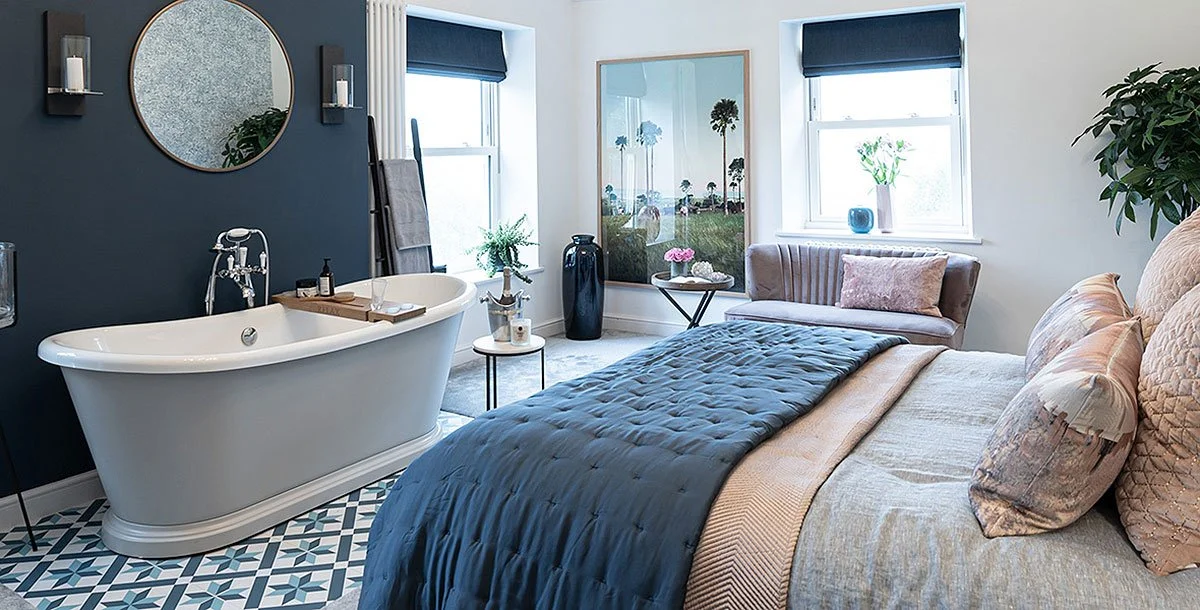 Modern bedroom with freestanding bathtub, blue bedding, decorative throw pillows, round mirror, and plants. The room features a dark accent wall and large windows with black blinds.