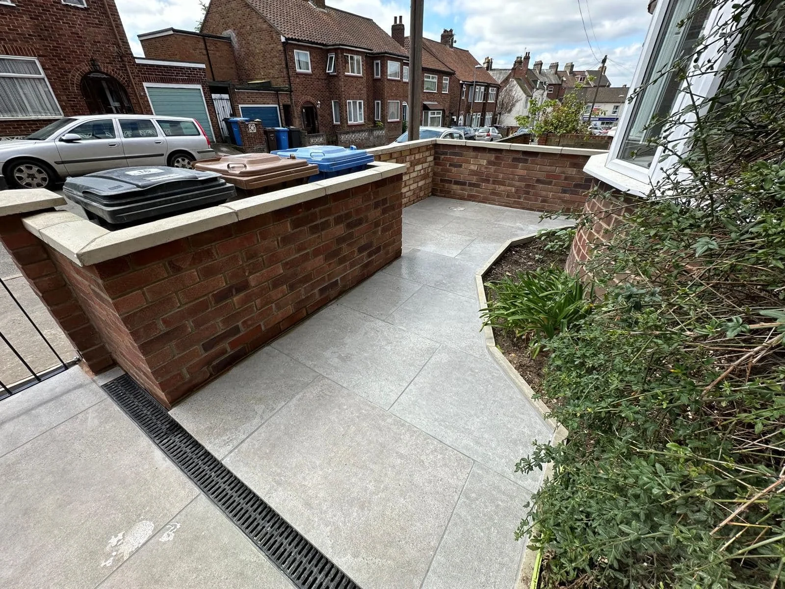 Residential front yard with brick walls, tiled pathway, and garden area; trash bins lined up; parked vehicles and houses in background.