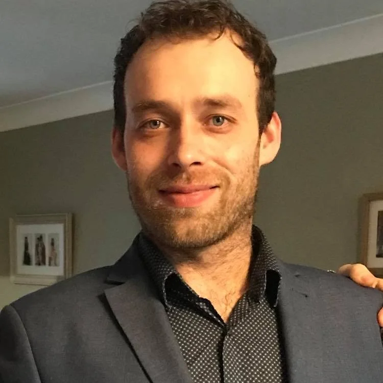 Man in a suit smiling indoors, with a light-colored wall and framed pictures in the background.