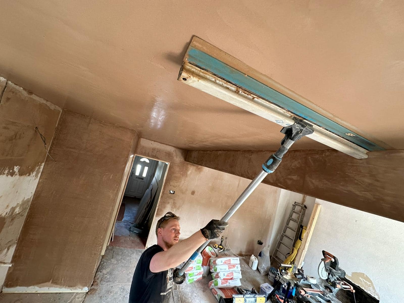 Man applying plaster on a ceiling with a trowel in a room under renovation, surrounded by construction tools and materials.