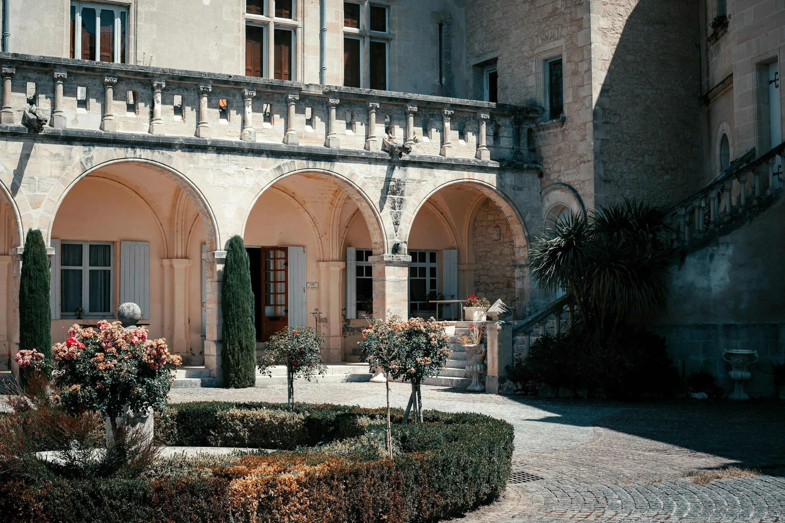 Exterior view of a historic stone building with arched entrances, a balcony, and lush landscaped garden with flowering bushes, cypress trees, potted plants, and a cobblestone pathway.