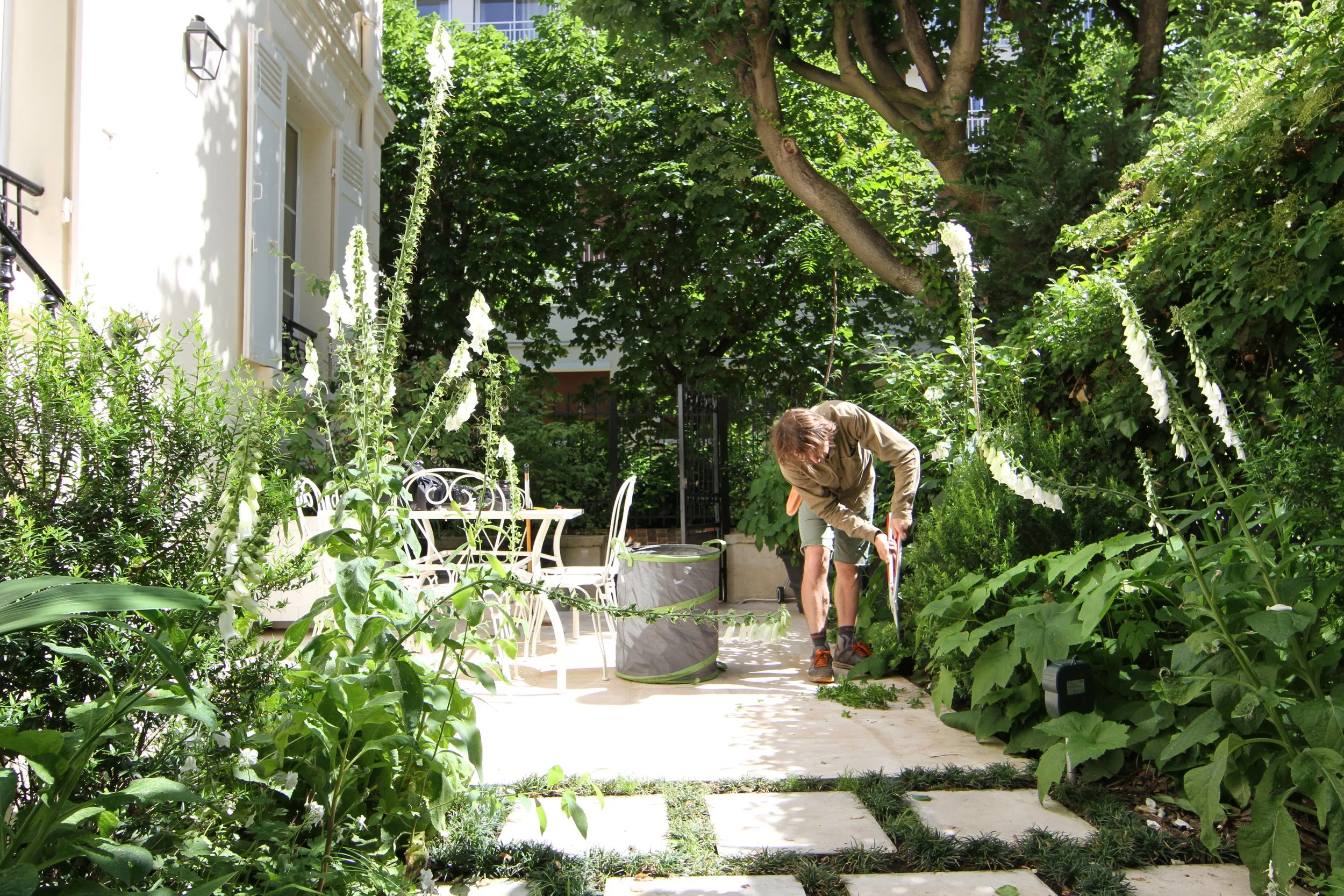 Un homme jardinant dans une zone de jardin verdoyante avec des arbres et des plantes, à côté d'une maison lumineuse avec des fenêtres et une porte noire.