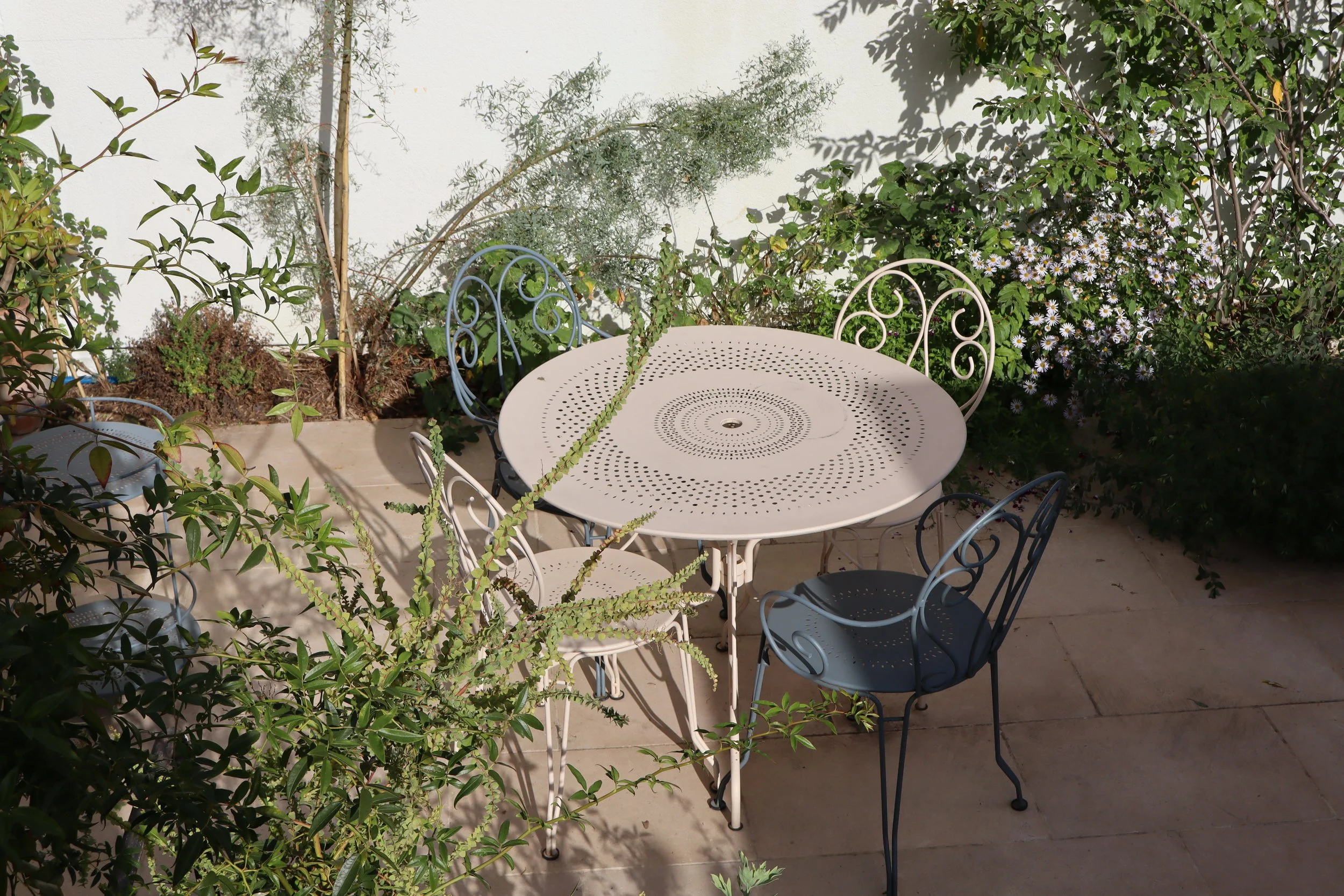 Coin repas de jardin aménagé par David Simonson avec table ronde perforée, chaises en fer forgé et massif fleuri