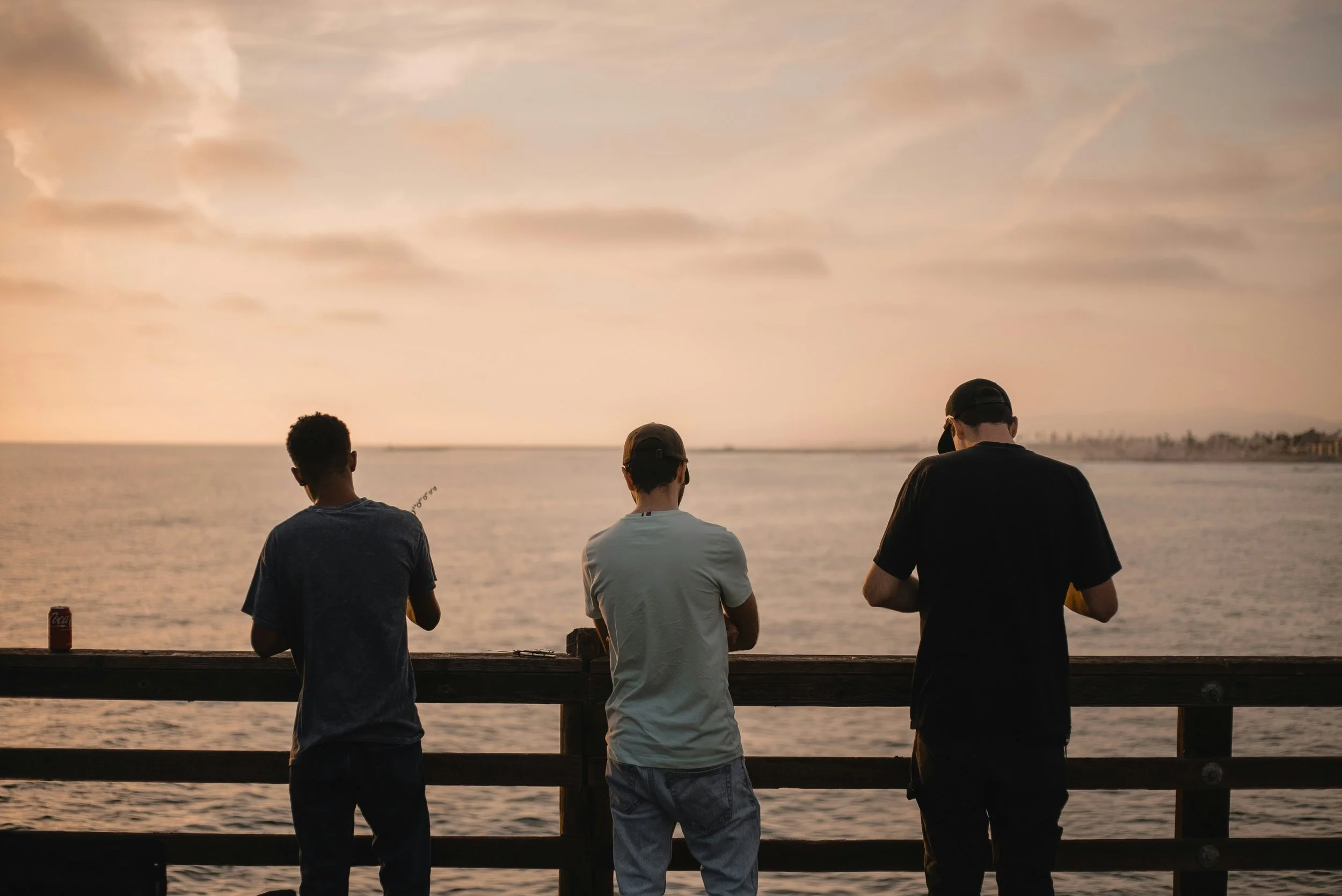 Three young men standing on a wooden pier by the water at sunset, with clouds and distant shoreline in the background.