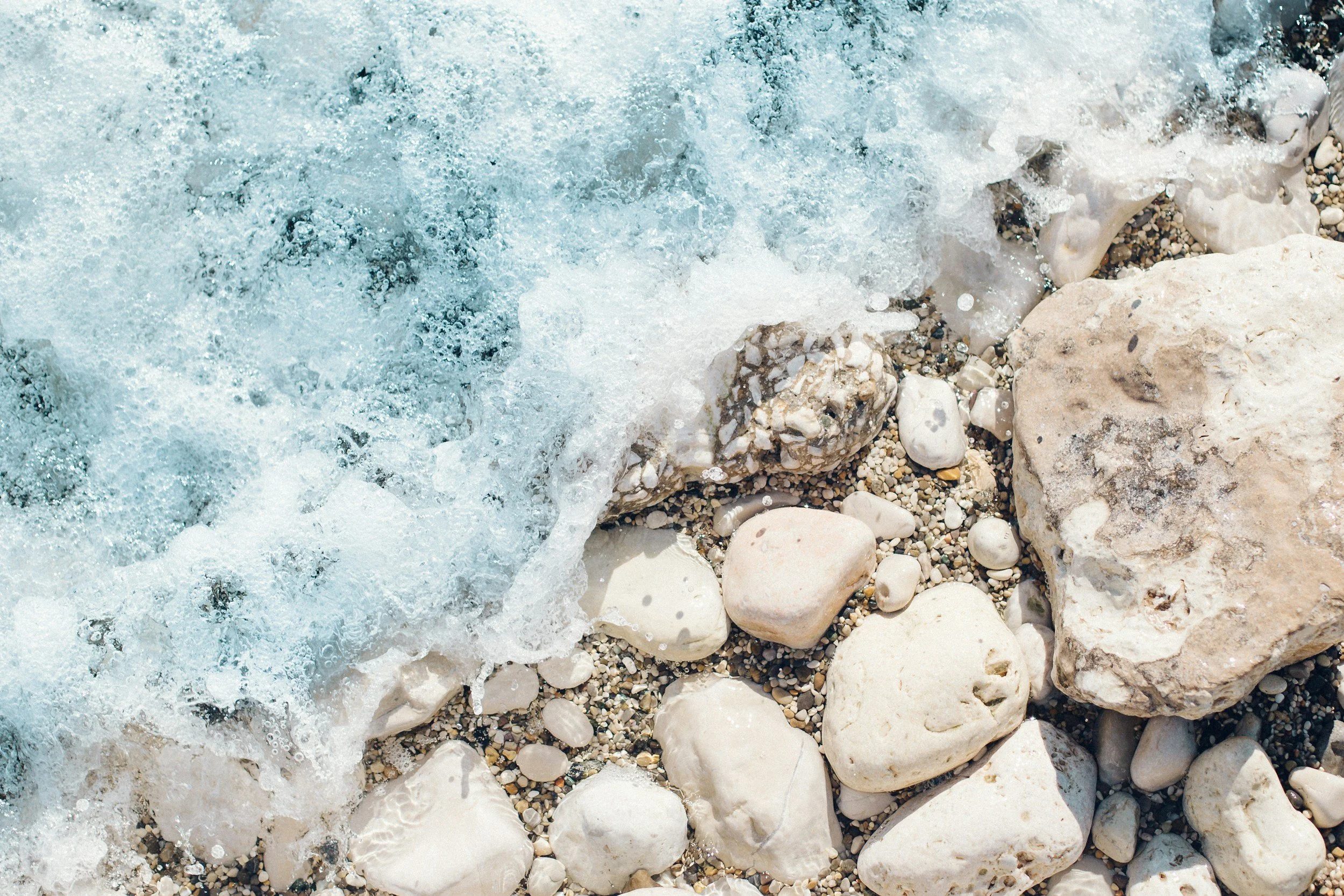 Close-up of a foamy ocean wave hitting a rocky shoreline with beige and white stones.