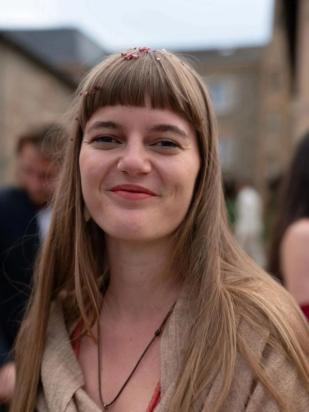 Close-up of a smiling woman with long, light brown hair, wearing a beige outfit and some red flower petals on her head, at an outdoor event.