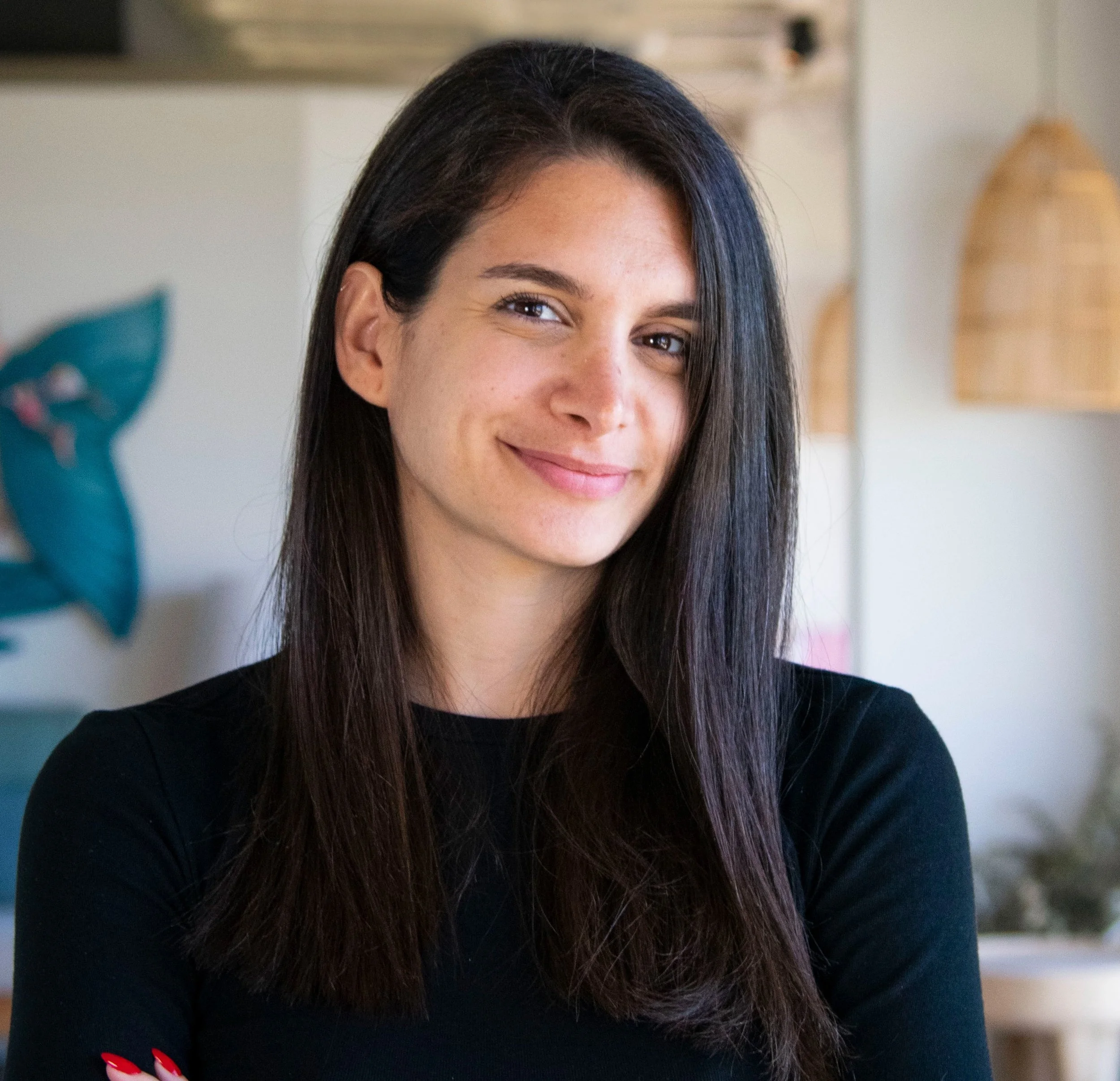 A young woman with long dark hair smiling at the camera in a bright room.