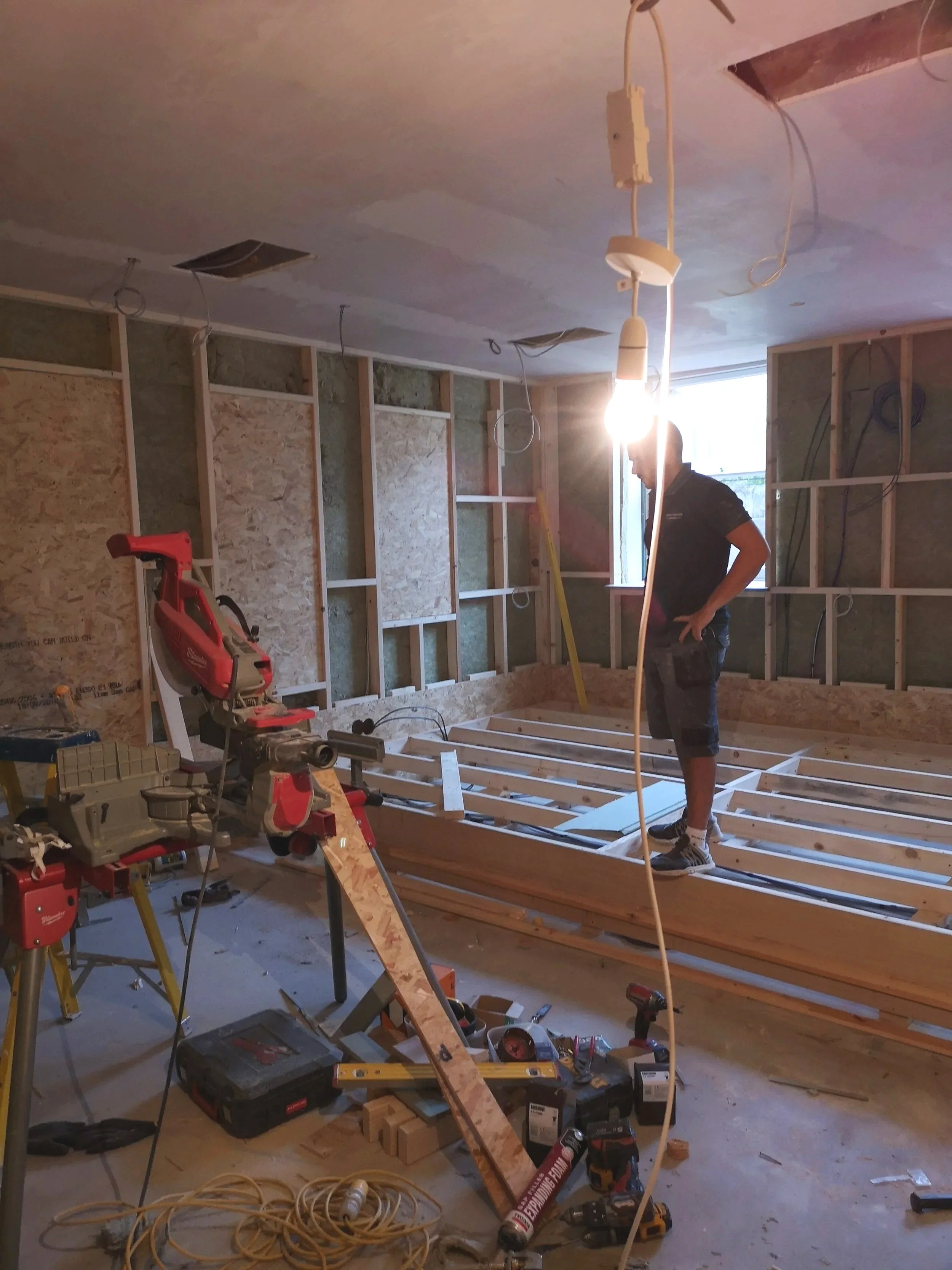 Construction worker standing on wooden floor framing in a room under renovation, with tools, equipment, and wiring scattered around, and a window in the background.