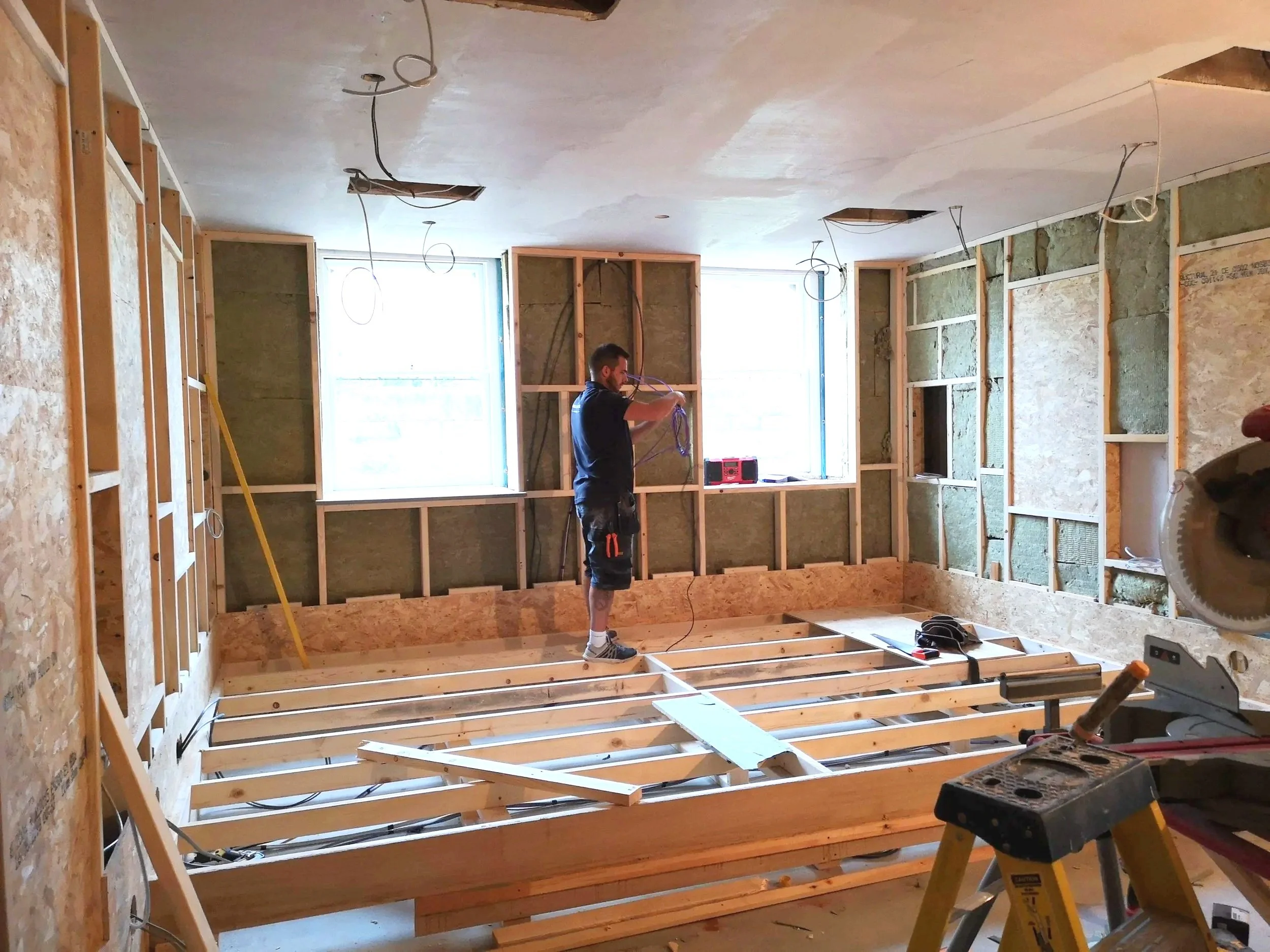 A construction worker standing on a wooden platform inside an unfinished room, working on electrical wiring. The room has two large windows, exposed wall studs with insulation, and a ceiling with cutouts for lighting fixtures. Construction tools and 