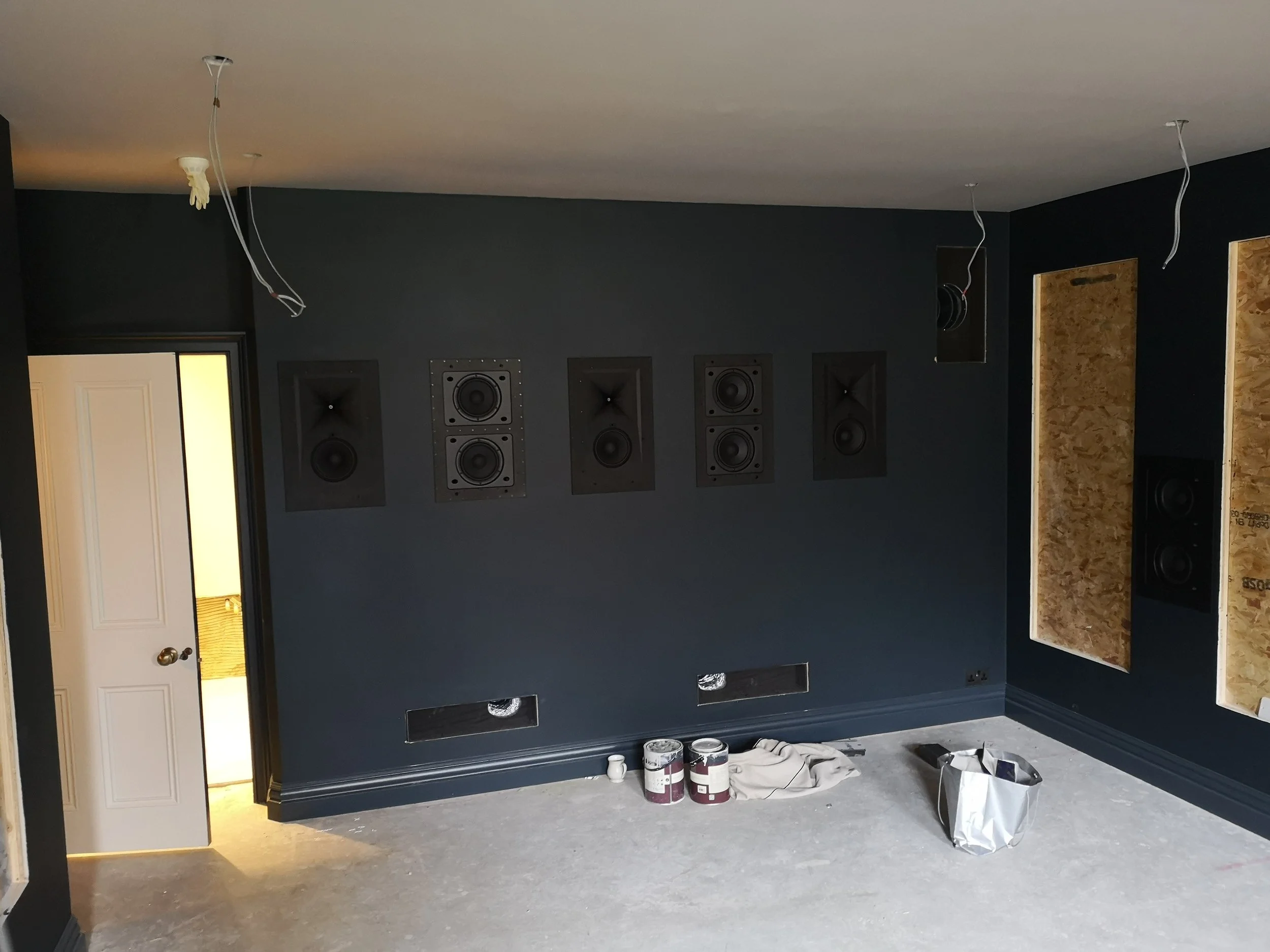 Room under renovation with dark blue walls, multiple black acoustic speakers mounted on the wall, unfinished wall panels, and construction materials on the floor.
