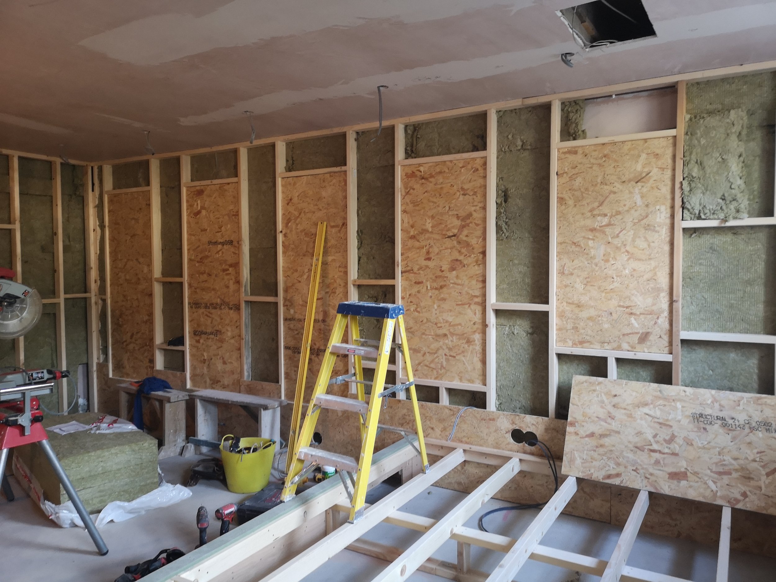 Interior of a room under construction, with exposed wooden framing, insulation, a yellow step ladder, and various tools and materials scattered on the floor.
