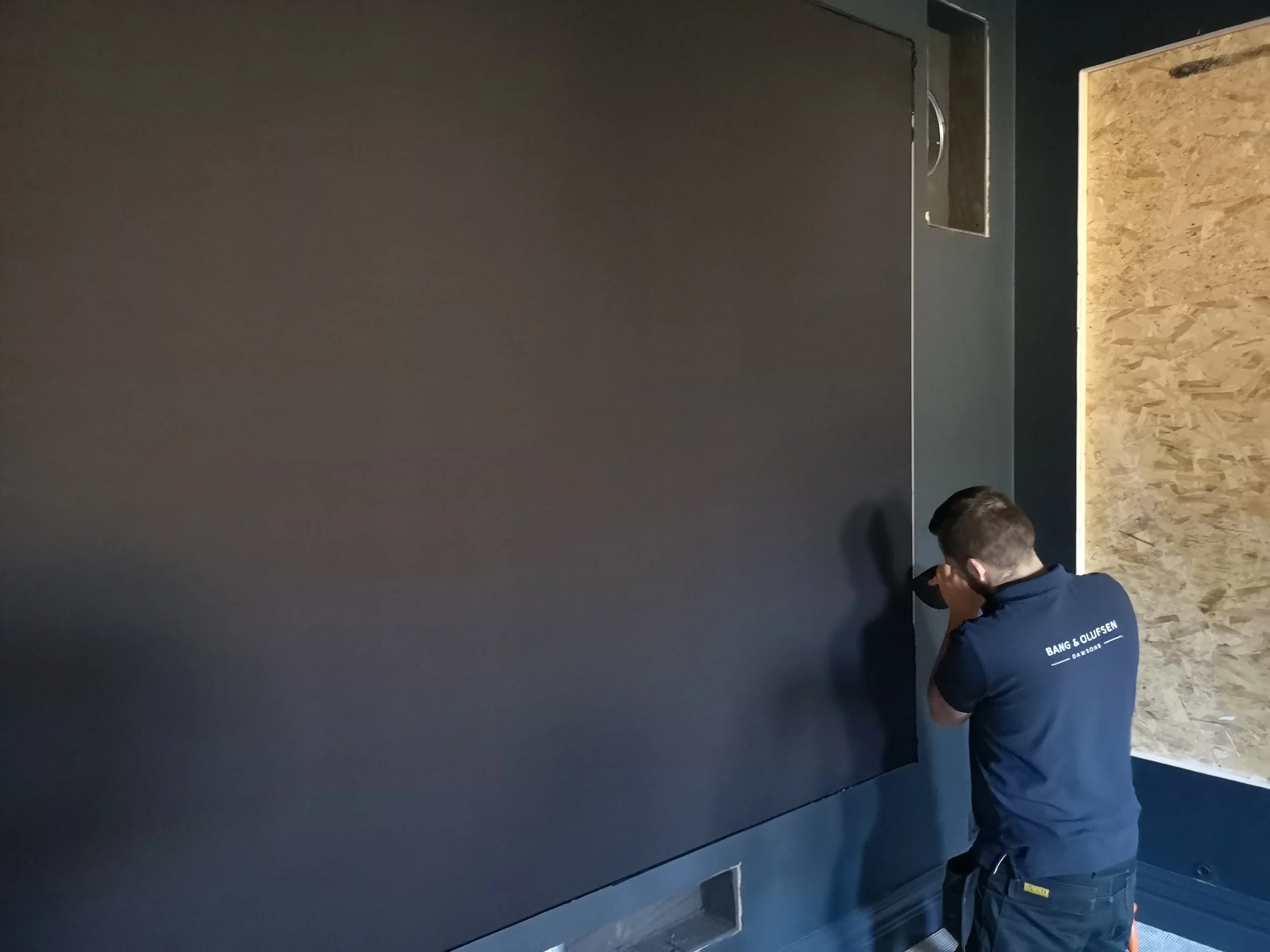 Two technicians install or repair a large flat-screen television on a dark wall. One technician is using a screwdriver, and the other is observing or assisting.
