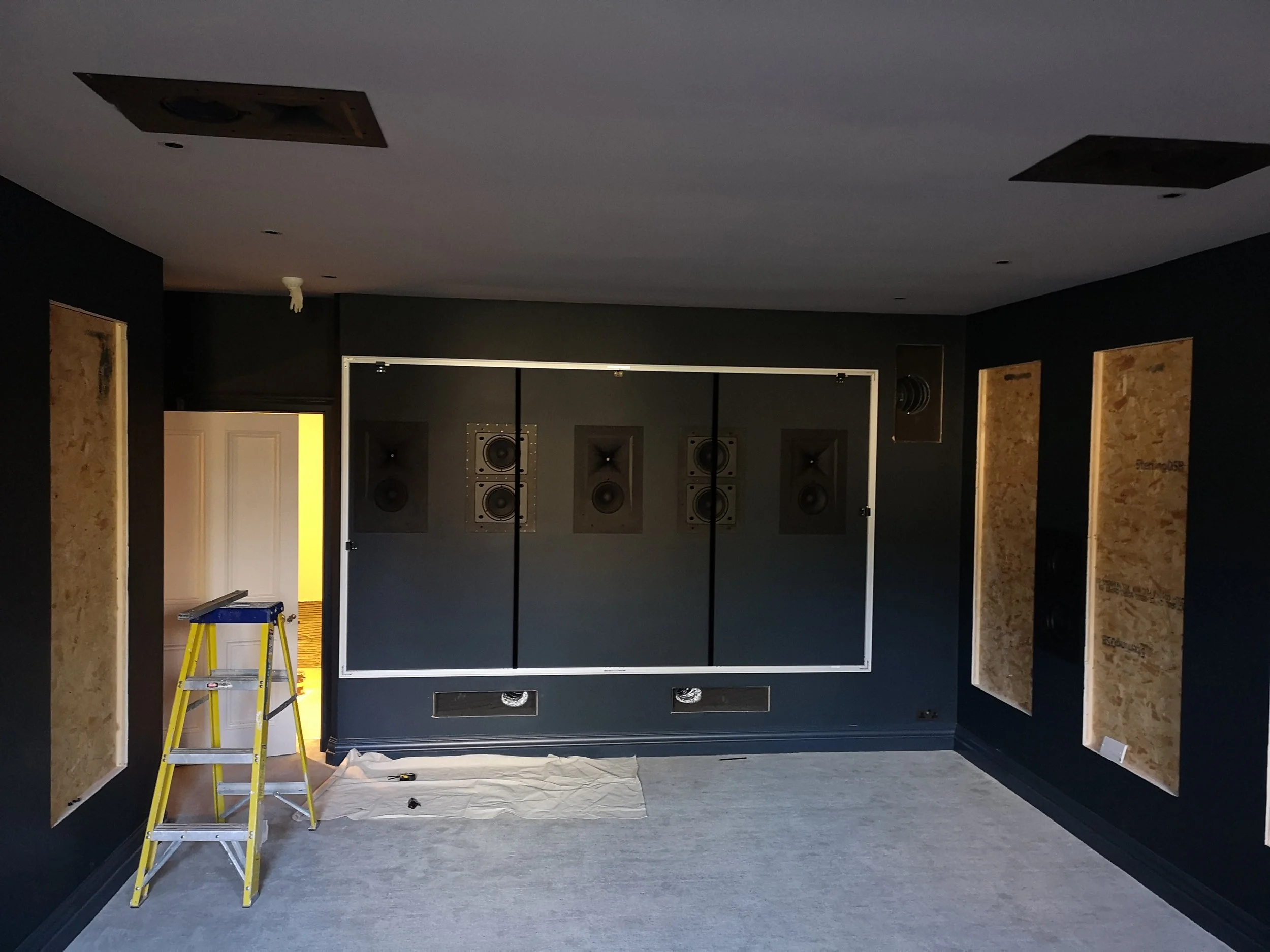 Room under renovation with black walls, soundproofing panels, and professional audio speakers installed on the back wall. A yellow step ladder and a cloth are on the floor, indicating ongoing work.