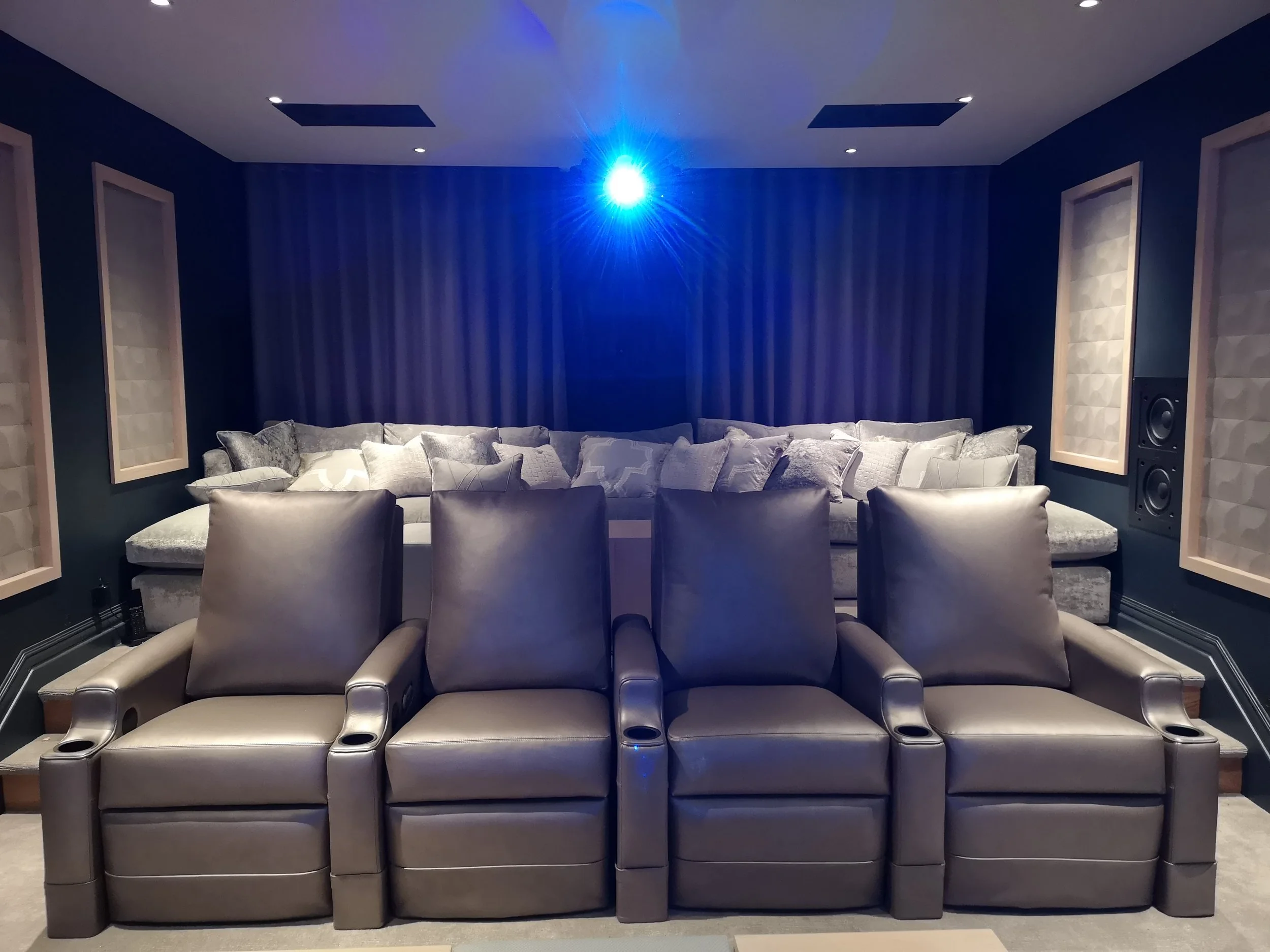 Home theater room with four leather recliner chairs in the foreground and a large sofa with numerous throw pillows in the background, blue walls, soundproofing panels, and a blue ceiling light.