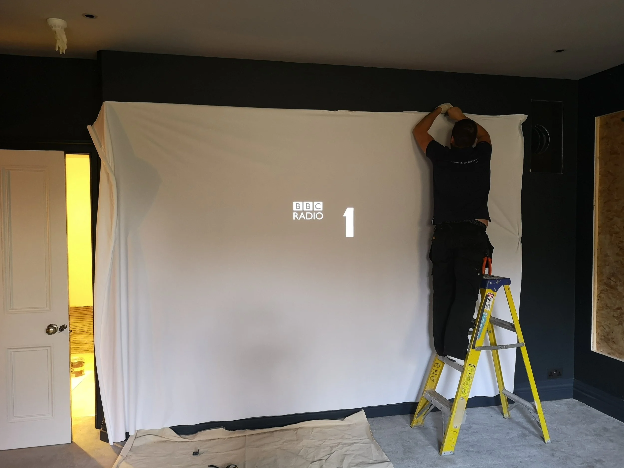 A man stands on a yellow ladder hanging a white fabric backdrop on a wall, with the BBC Radio 1 logo and number 1 displayed on the fabric.