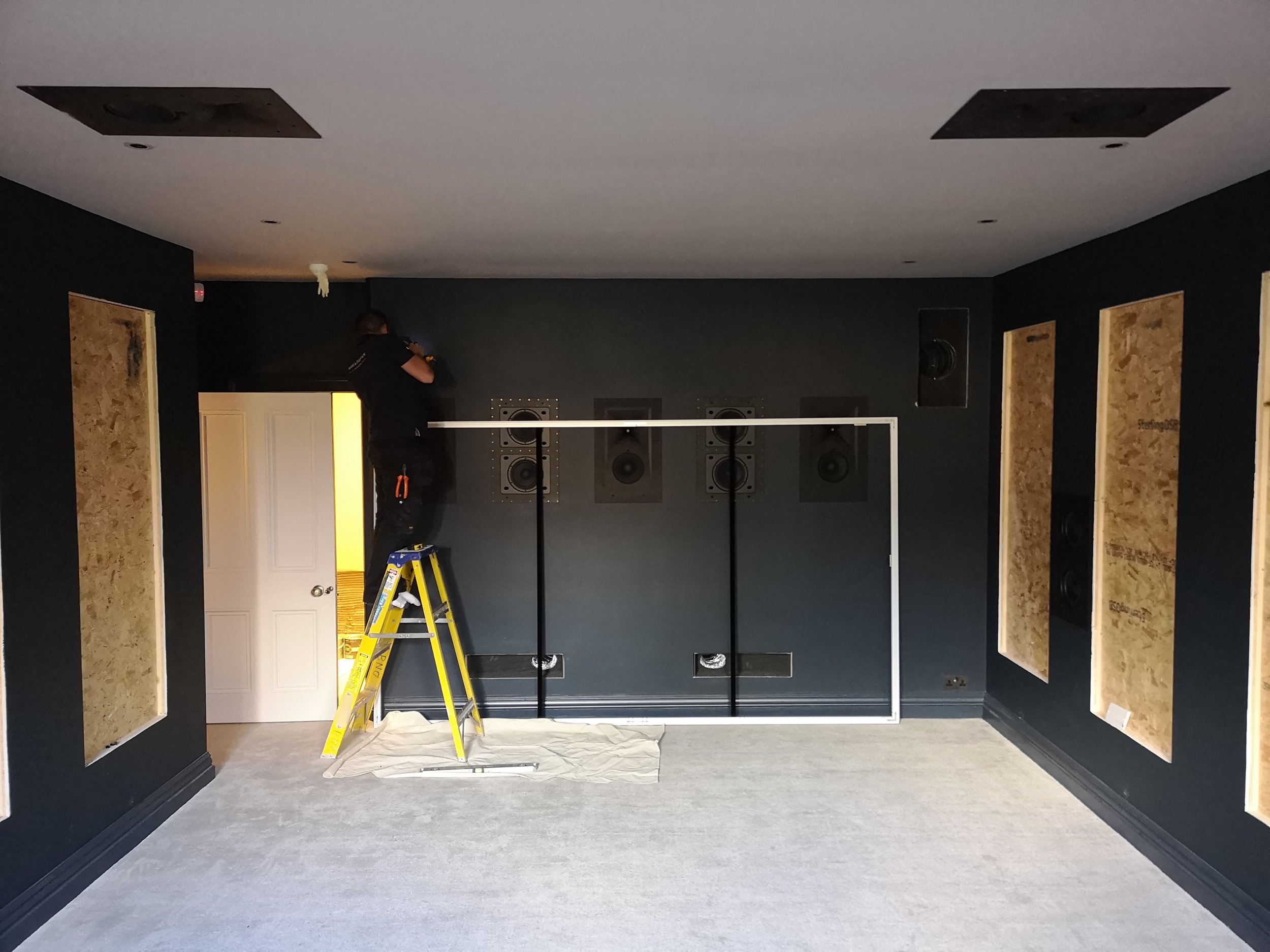 A man on a ladder installing or adjusting sound speakers in a dark blue room with acoustic panels on the walls. A white frame is placed on the floor in front of him, and there are ventilation vents and an open door leading to a yellow-lit room.