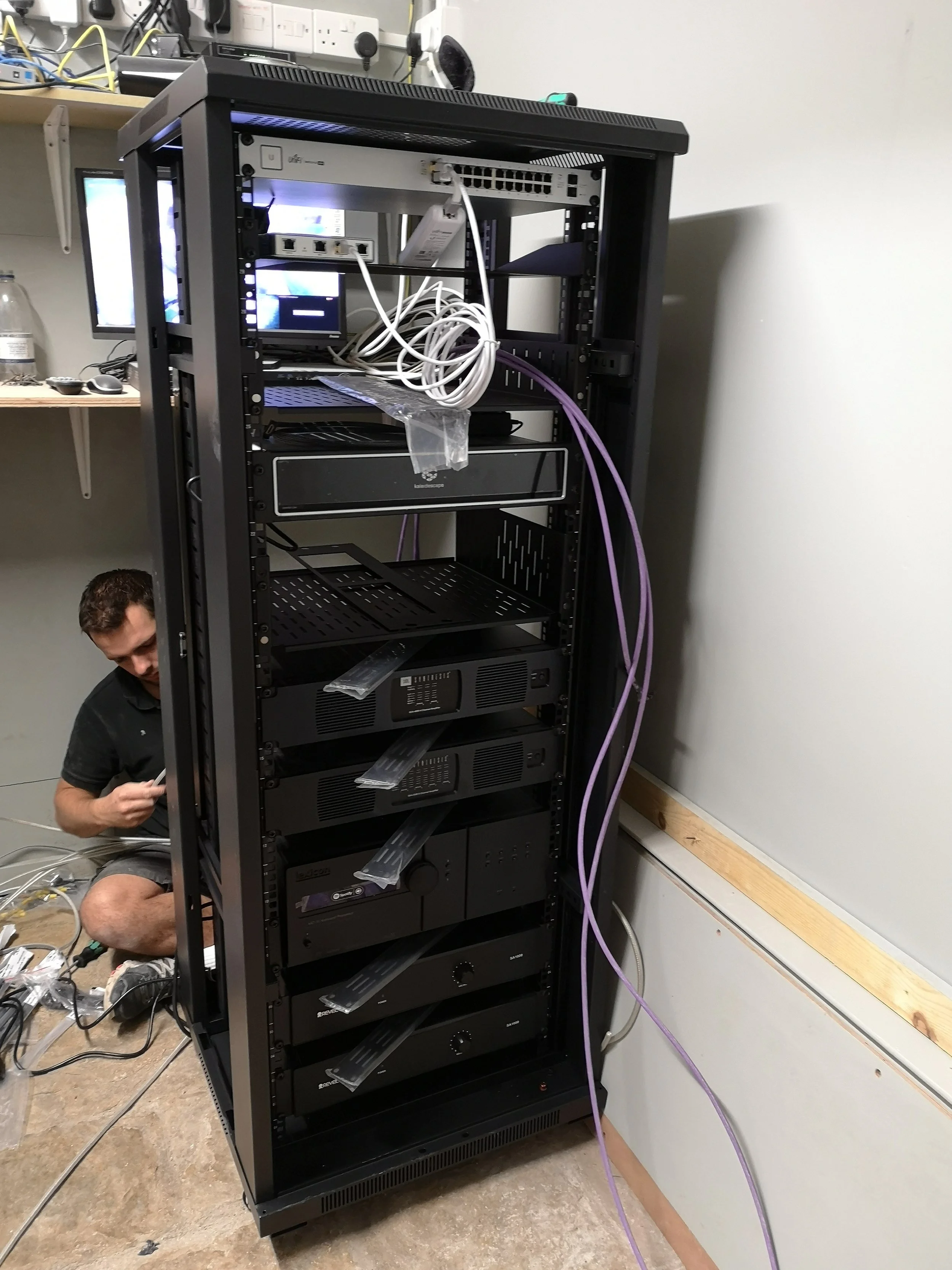 A person working on a server rack with multiple servers and networking equipment mounted, wires connecting the devices, and a workspace with shelves and tools nearby.
