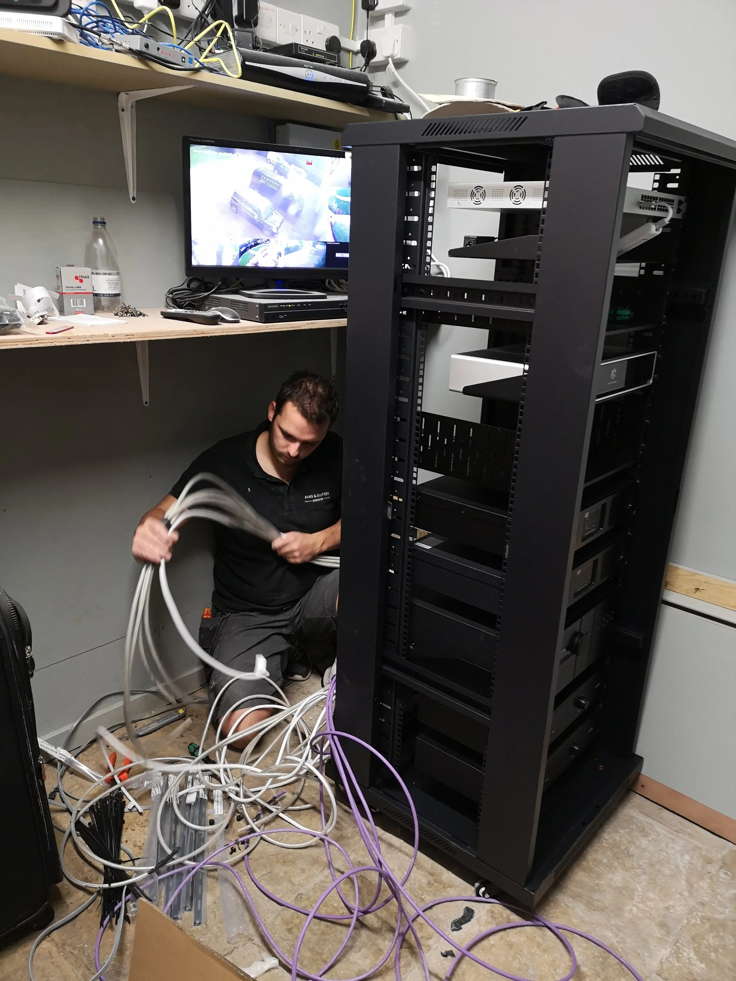 A technician working on a server rack with multiple cables, in a room with a workbench, a monitor, and networking equipment.
