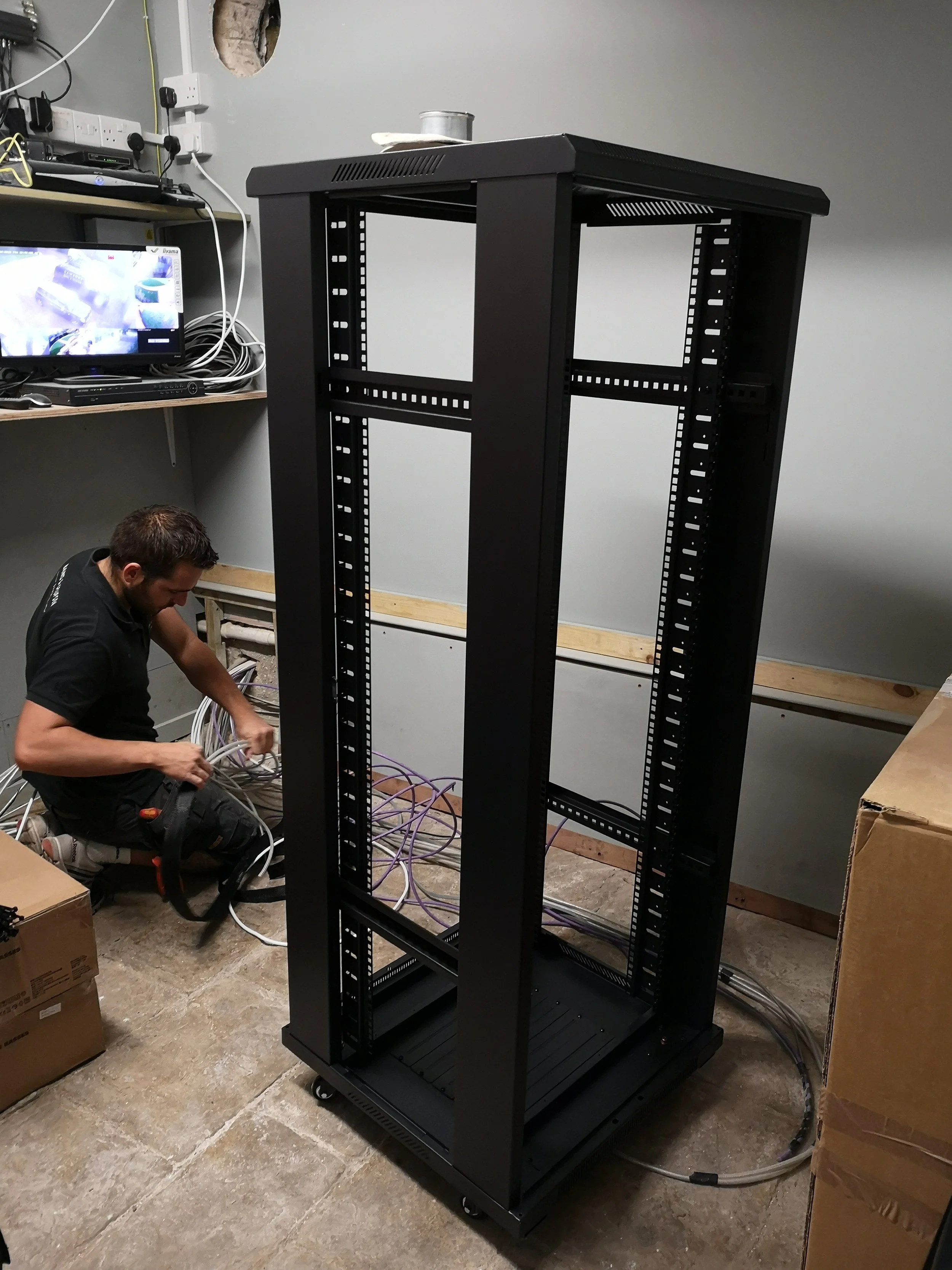 A man installing or working on a black server rack in a room with cables and boxes around.