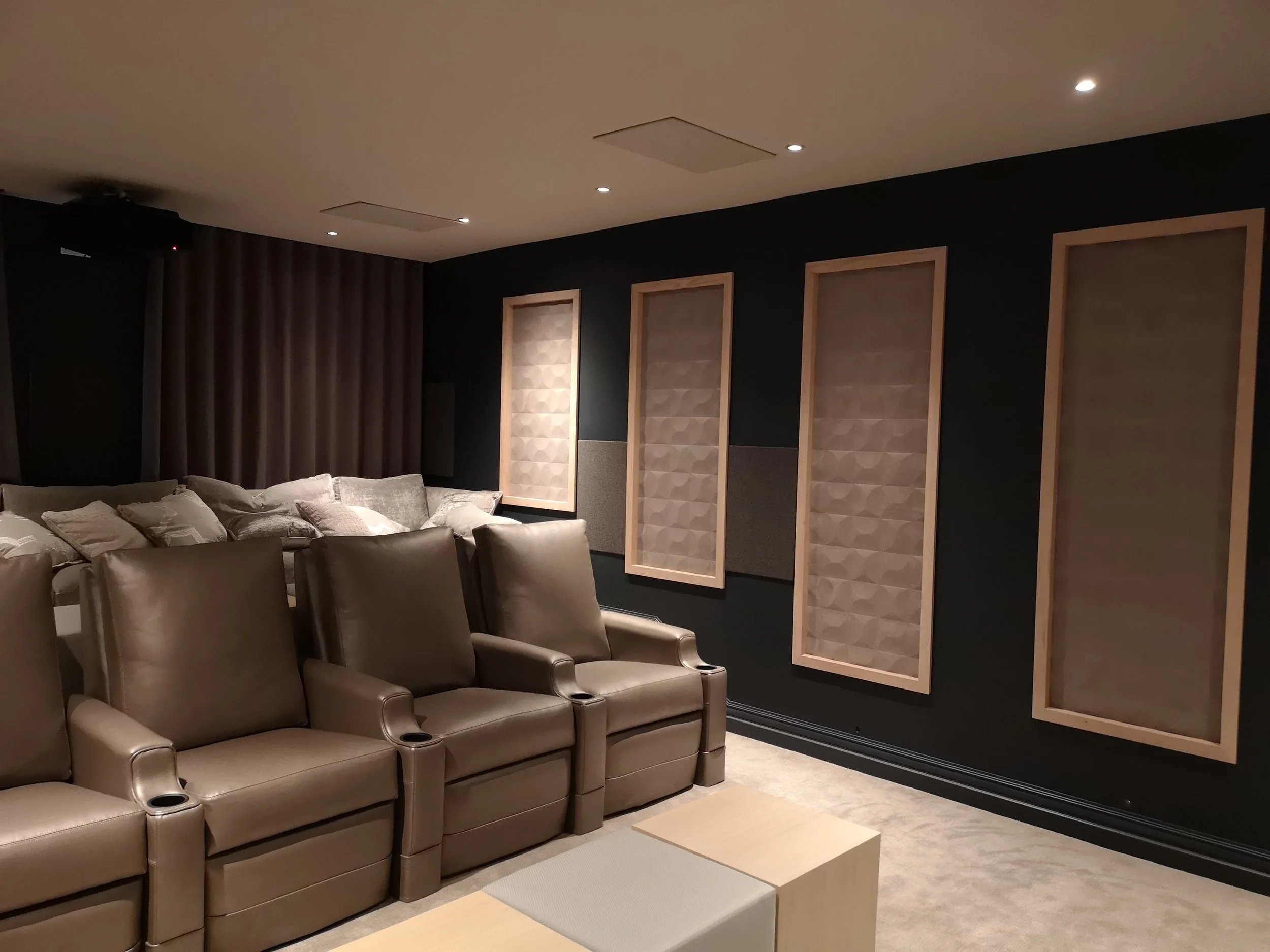 A modern home theater room with brown leather chairs, a beige ottoman, and acoustic wall panels on a black wall.