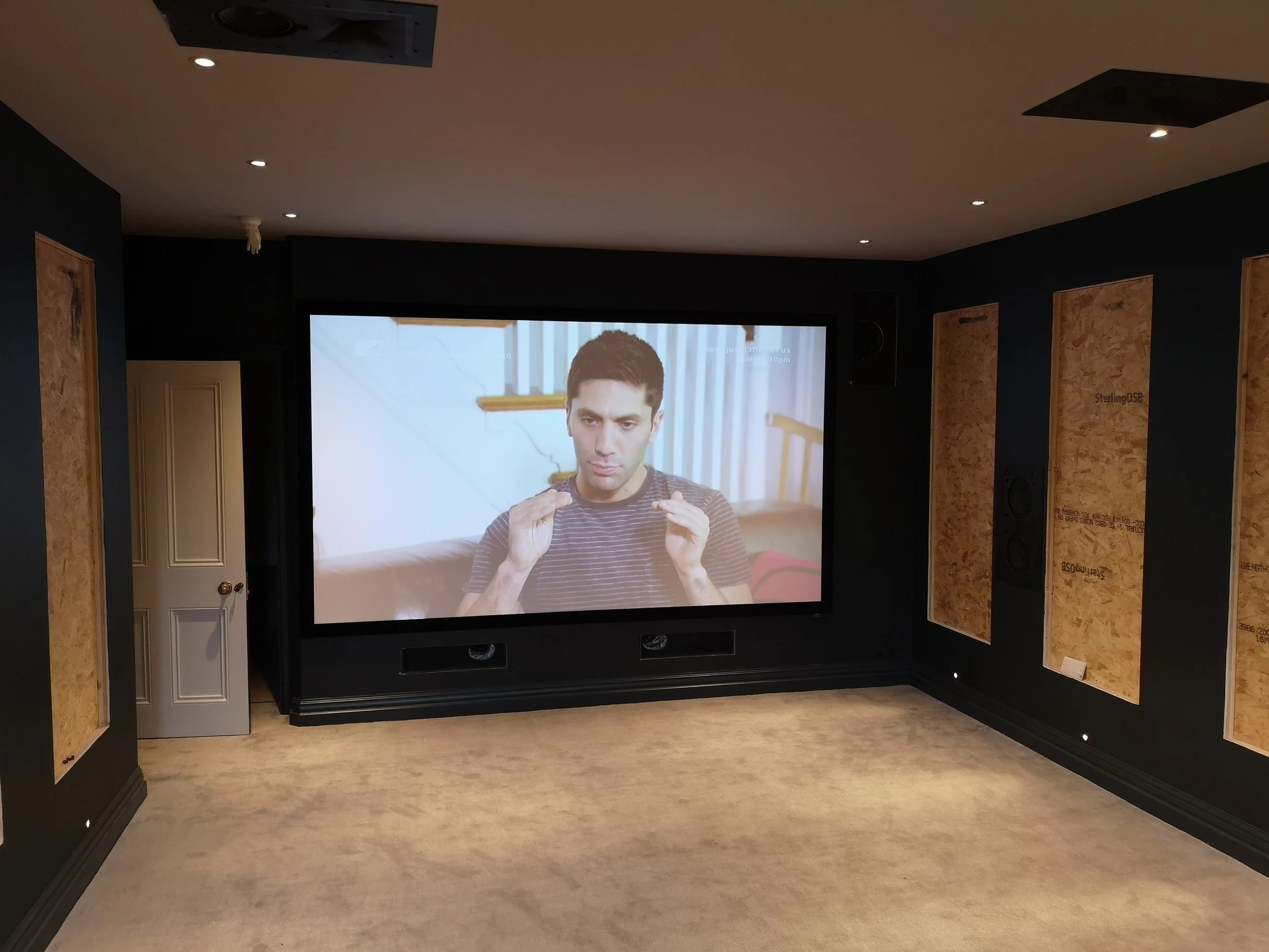 Empty home theater room with a large projection screen showing a man talking. The walls are partially unfinished with wooden panels, and the room has recessed ceiling lights and a beige carpet.