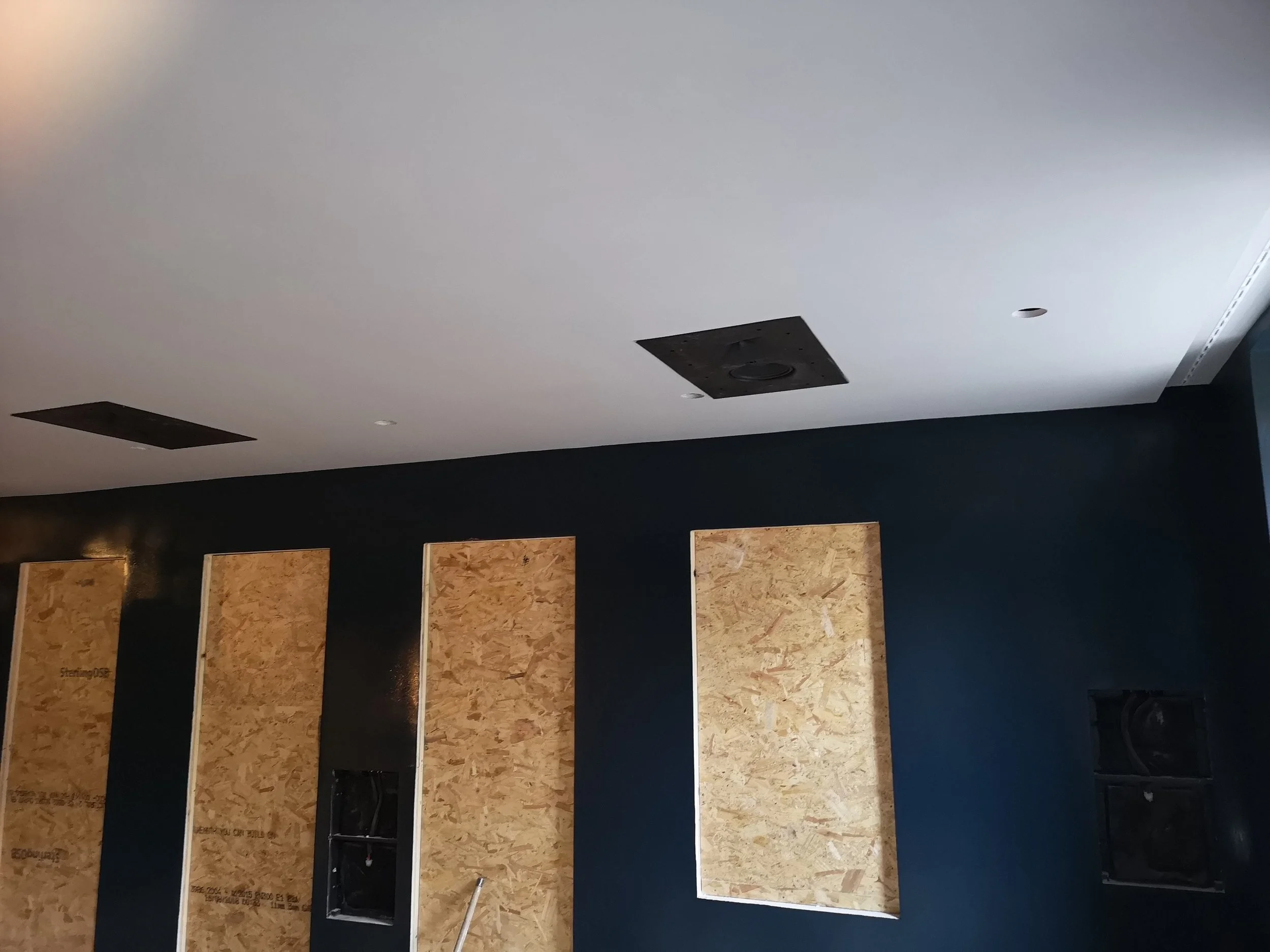 Interior ceiling with black speakers installed, wall with wooden panels and black speaker boxes, and a section of dark wall paint.