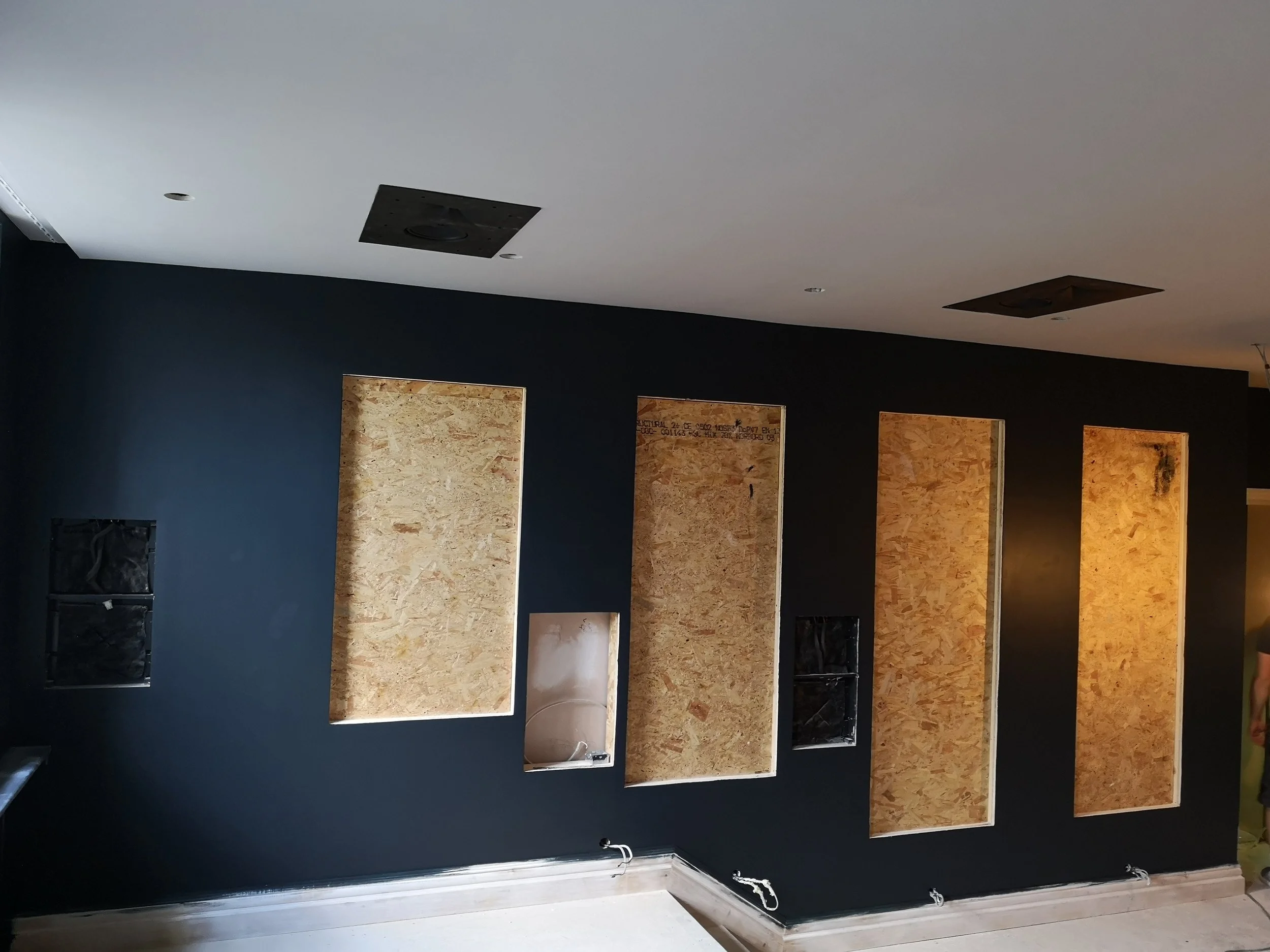 Interior wall with four rectangular cutouts framed with plywood, an open electrical box, and a small sink with plumbing in the middle cutout, all against a dark-colored wall.