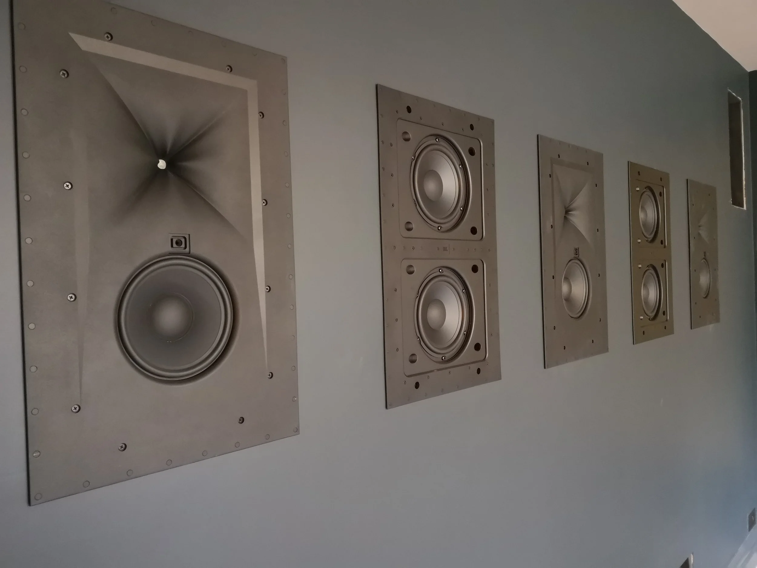Four metallic wall-mounted speakers arranged in a row on a room wall.