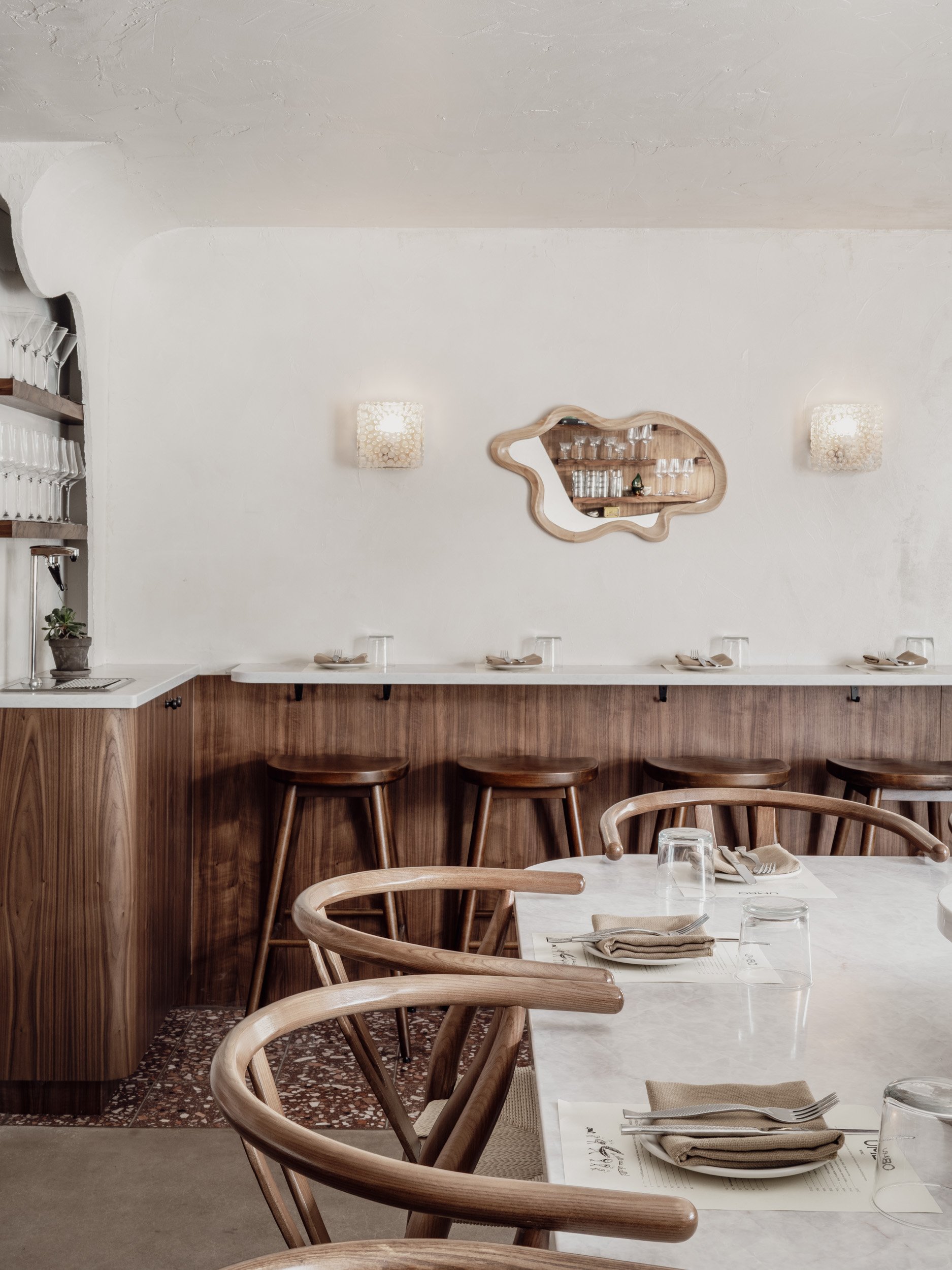 Interior of restaurant dining space with counter stools, warm woods, and soft ceiling textures.