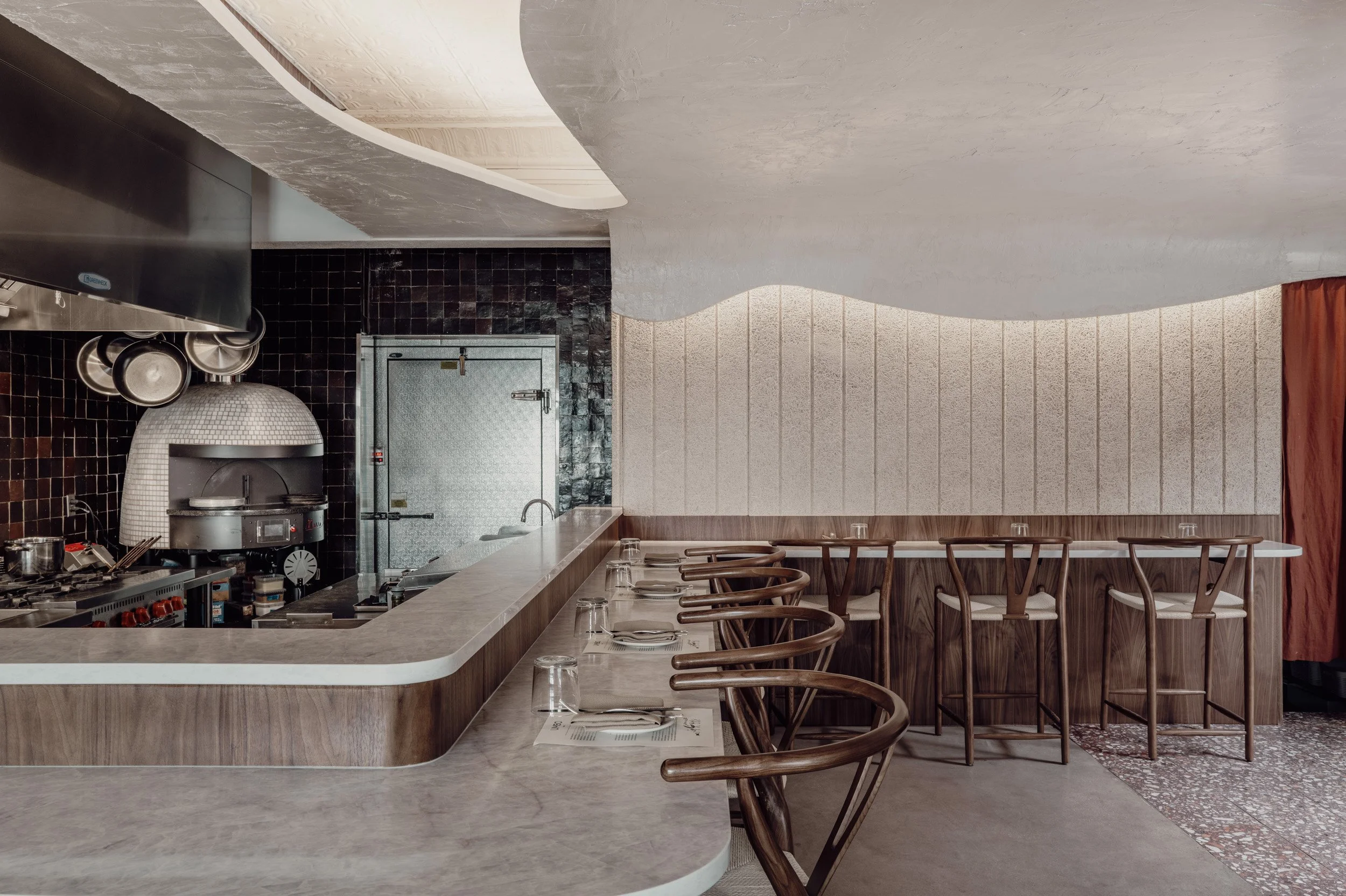 Interior of restaurant dining space with counter stools, warm woods, and soft ceiling textures.