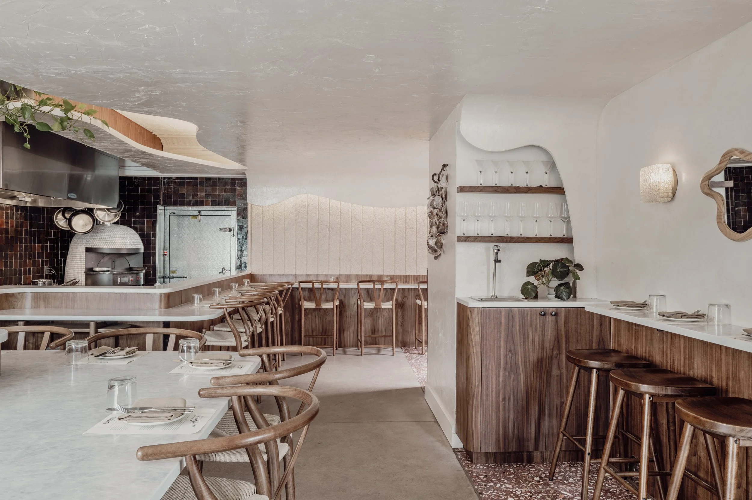 Interior of restaurant dining space with counter stools, warm woods, and soft ceiling textures.
